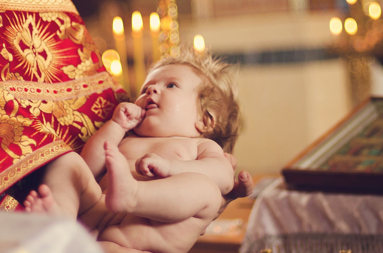 Niemowlę w ramionach osoby w bogato zdobionej szacie liturgicznej, prawdopodobnie podczas ceremonii chrztu, w tle świece i ikona.