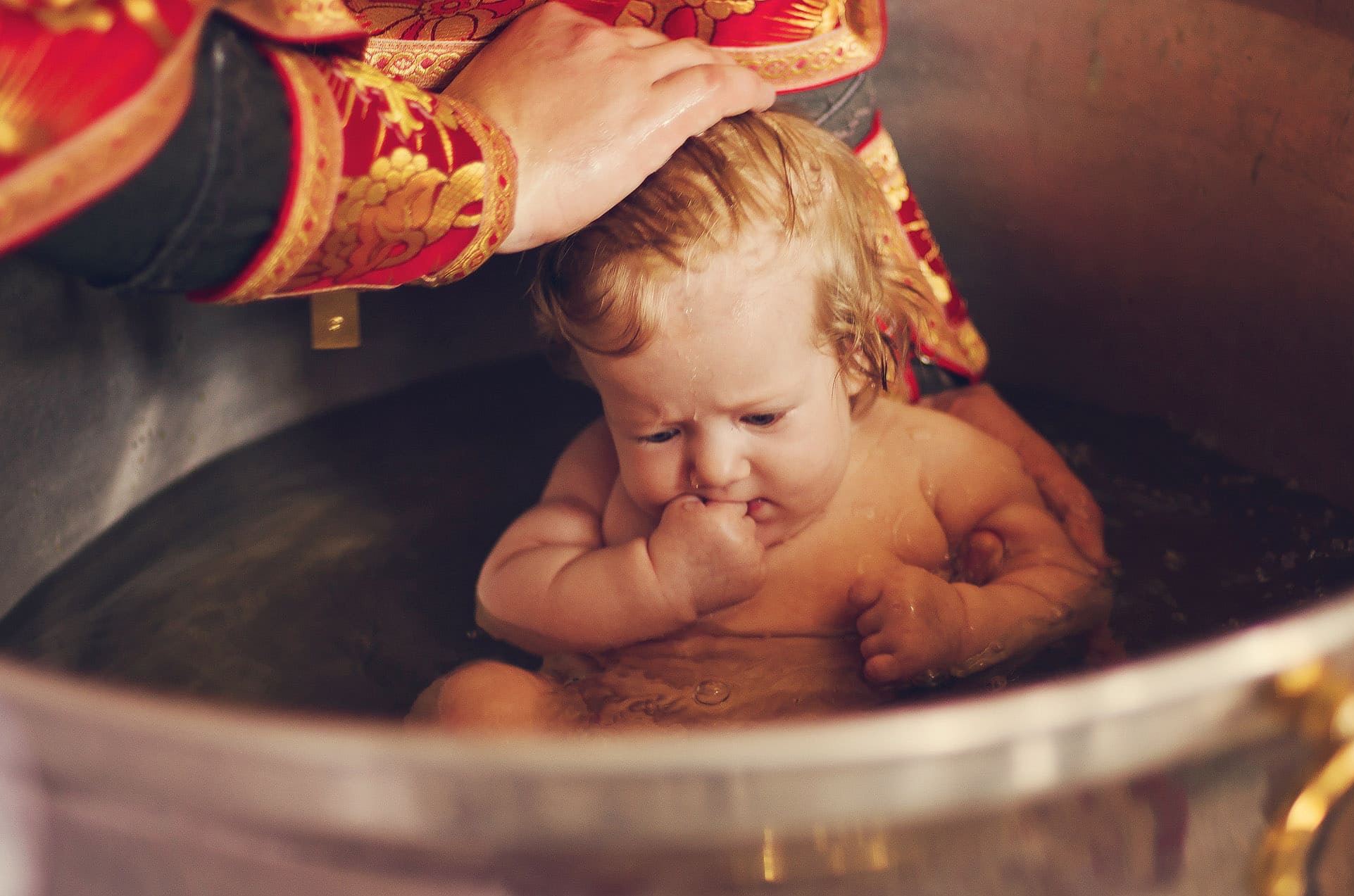 Dziecko w wanience podczas chrztu, woda spływa po jego twarzy, dłoń kapłana w bogato zdobionym ornacie dotyka głowy dziecka. Delikatne światło podkreśla emocje.