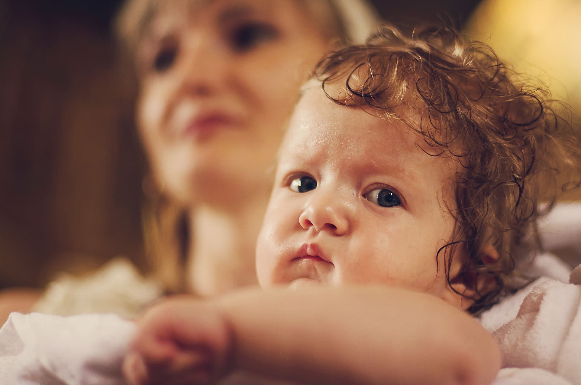 Portret niemowlęcia z kręconymi włosami na tle rozmytej sylwetki kobiety, uchwycony w ciepłym oświetleniu, z bliska, z delikatnym uśmiechem na twarzy dziecka.