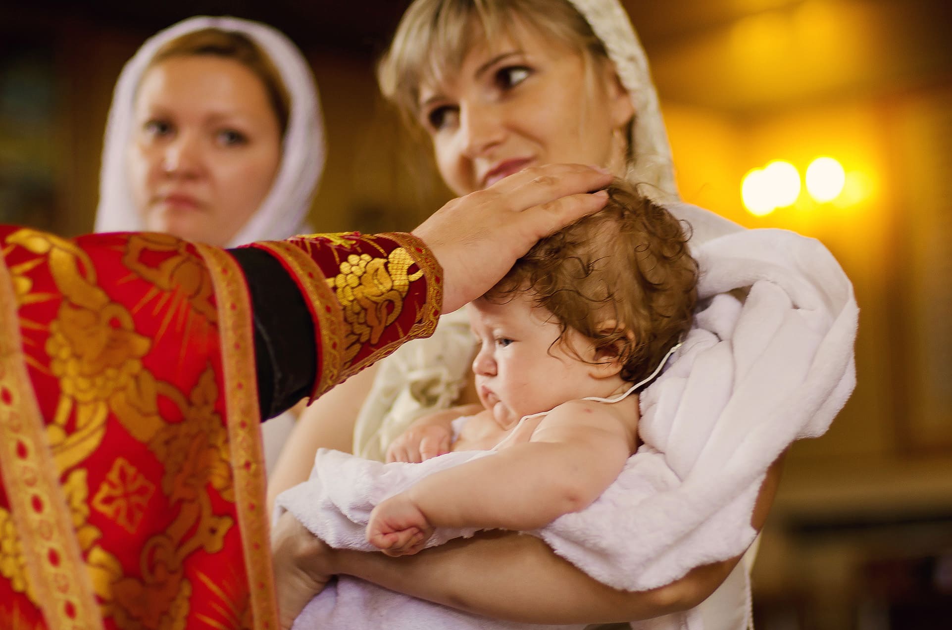 Chrzest dziecka w cerkwi: matka trzyma niemowlę owinięte w biały ręcznik, ksiądz w bogato zdobionej szacie błogosławi, w tle kobieta w chuście.