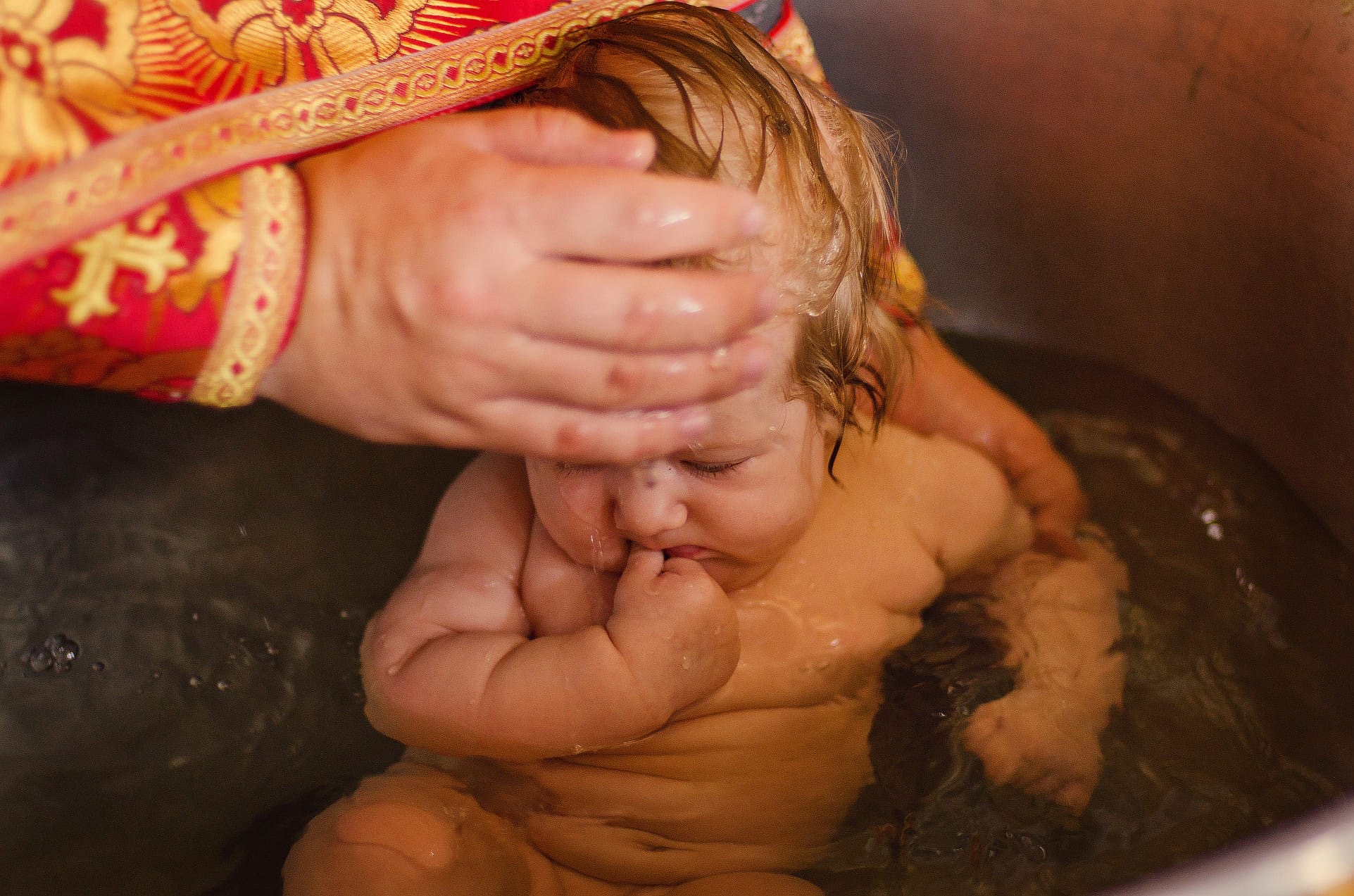 Kąpiel noworodka podczas ceremonii chrztu, woda spływa po twarzy dziecka, dłoń kapłana w bogato zdobionej szacie. Ujęcie z góry, ciepłe światło.