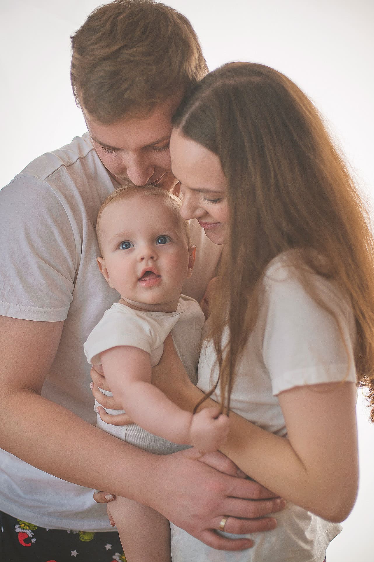 Rodzinne ujęcie w Lublinie: Mama i tata tulą niemowlę w białym body. Delikatne światło podkreśla bliskość i miłość. Uśmiechnięte twarze rodziców.