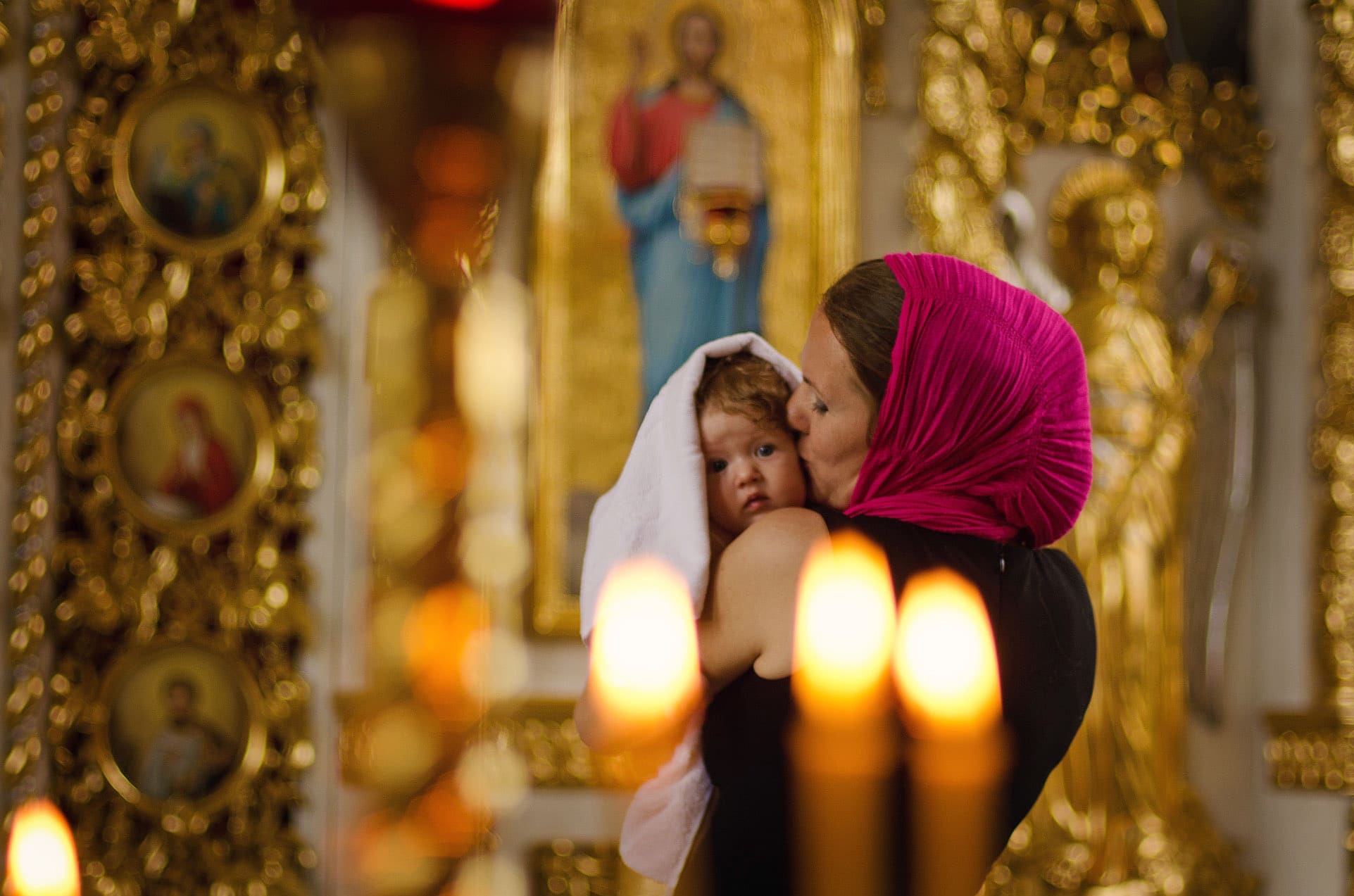 Matka w różowej chuście całuje w głowę niemowlę owinięte białym ręcznikiem na tle bogato zdobionego wnętrza cerkwi, z widocznymi ikonami i płonącymi świecami.