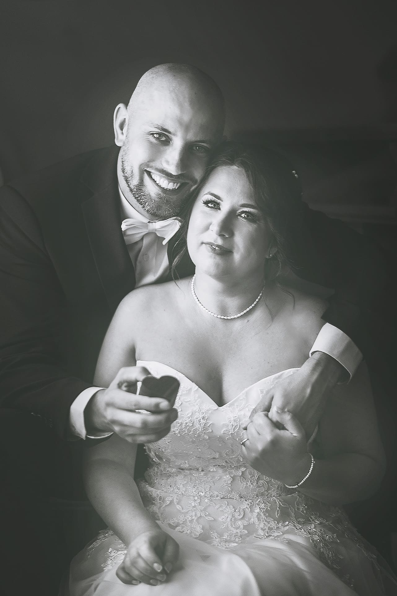 Czarno-białe, portretowe ujęcie uśmiechniętej pary w strojach ślubnych. Pan obejmuje Panią, trzymając w dłoniach małe, sercowate pudełko. Romantyczna atmosfera.