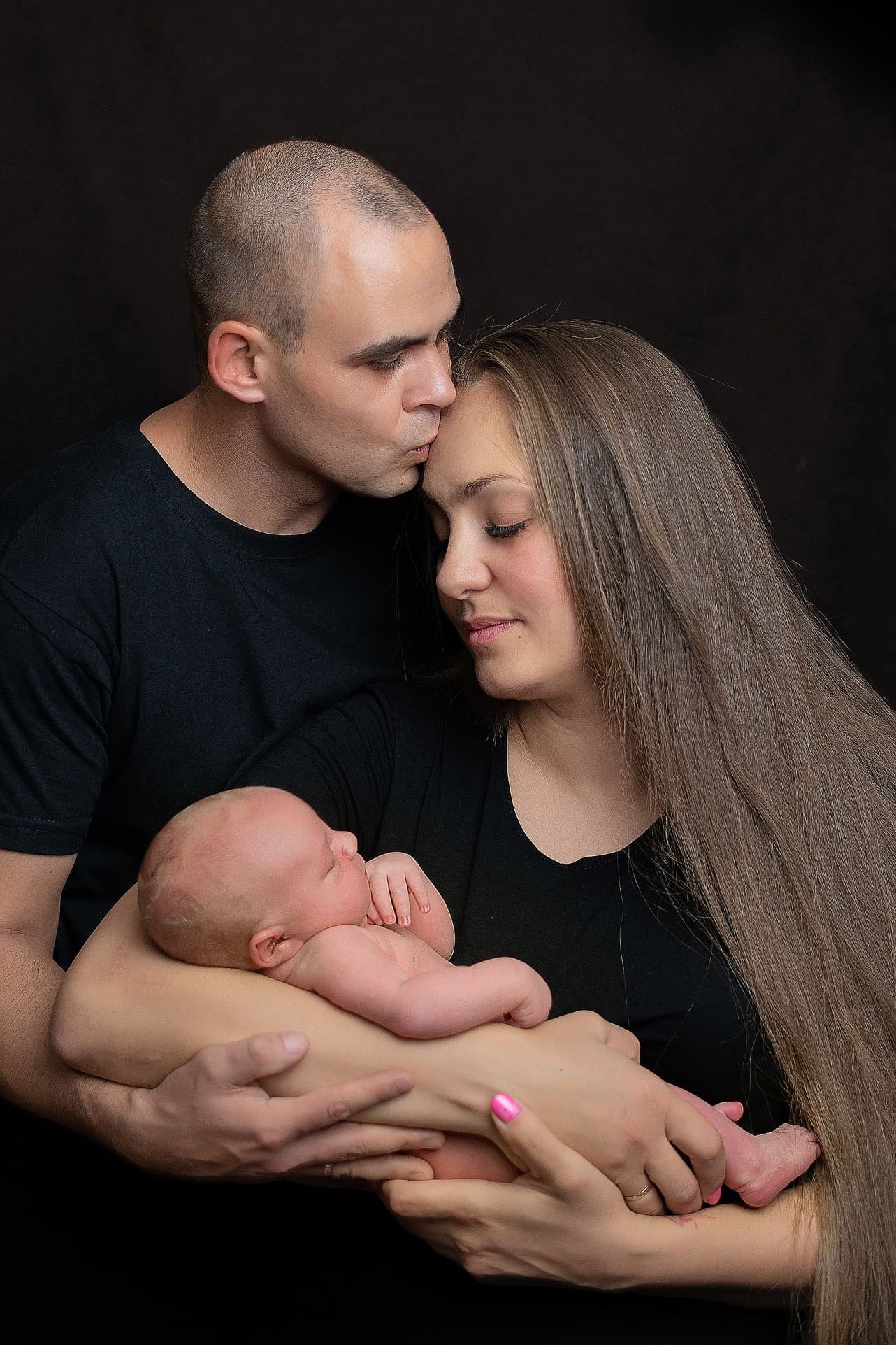 Rodzinne ujęcie: ojciec całuje matkę w czoło, trzymając śpiącego noworodka na rękach. Ciemne tło, portret w bliskim planie, skupienie na emocjach i więzi.