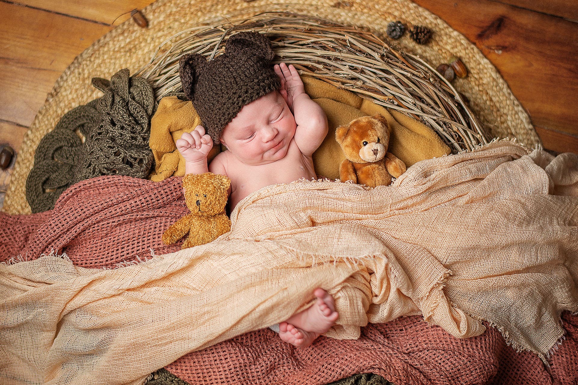 Uśmiechnięty noworodek w brązowej czapce z uszami misia, leżący w koszu z pluszowymi misiami i miękkimi tkaninami w ciepłych odcieniach brązu i beżu.