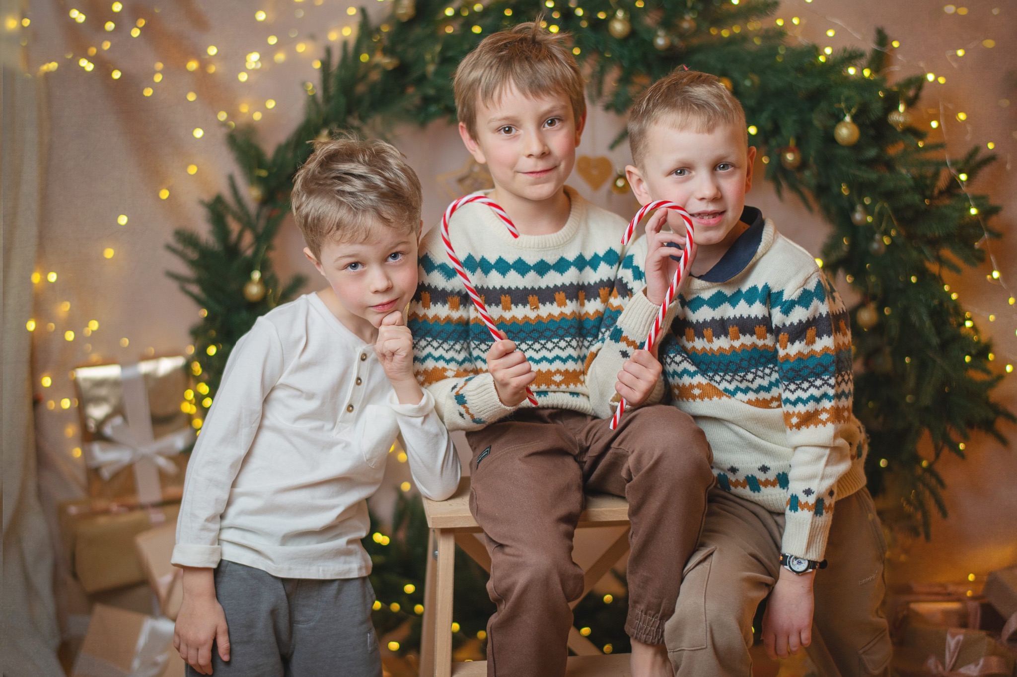 Trzech chłopców w świątecznych swetrach pozuje z lizakami na tle choinki i prezentów, ciepłe światło, rodzinna atmosfera, sesja w studio.