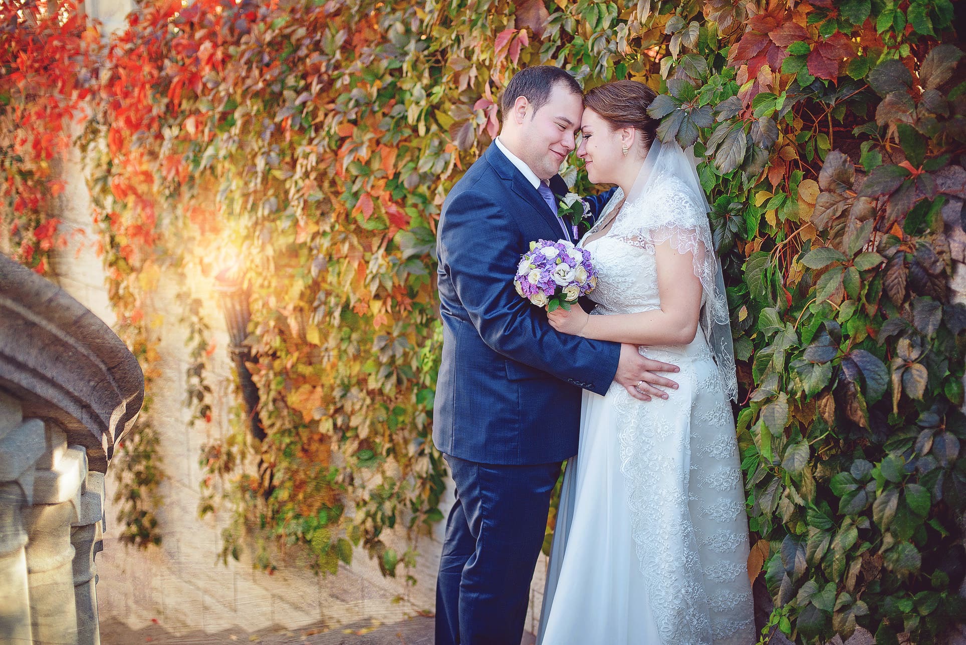 Para młoda w objęciach na tle ściany z bluszczu w jesiennych barwach. Panna młoda w koronkowej sukni trzyma bukiet. Pan młody w granatowym garniturze.