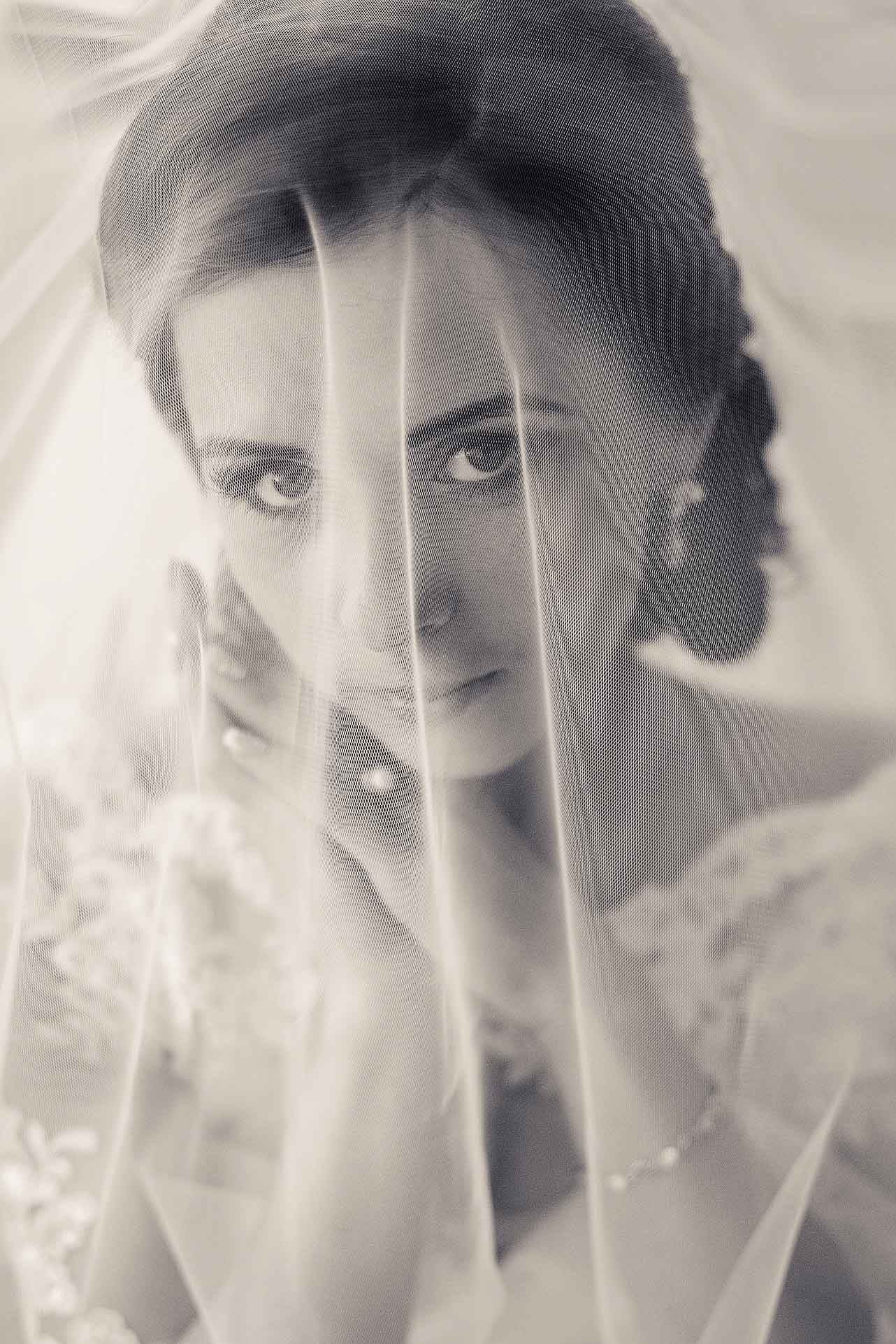 Portret panny młodej w sepii, z delikatnym welonem na twarzy, podkreślającym jej spojrzenie. Subtelna biżuteria dodaje elegancji. Ujęcie z bliska, skupione na emocjach.