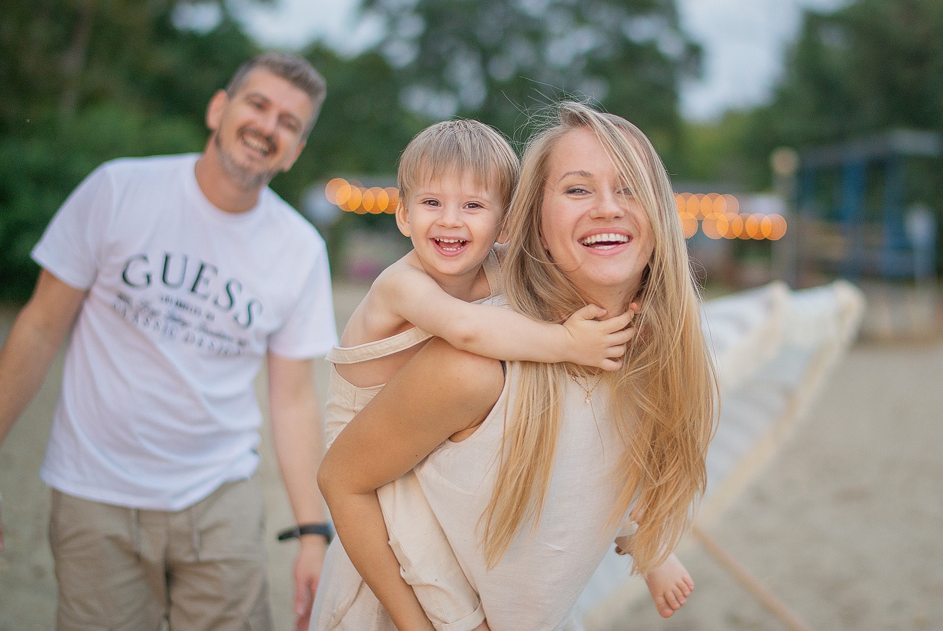 Radosna sesja rodzinna na plaży: uśmiechnięta mama z synkiem na barana, tata w tle. Naturalne światło, ciepłe kolory, wakacyjna atmosfera. Idealne na pamiątkę.