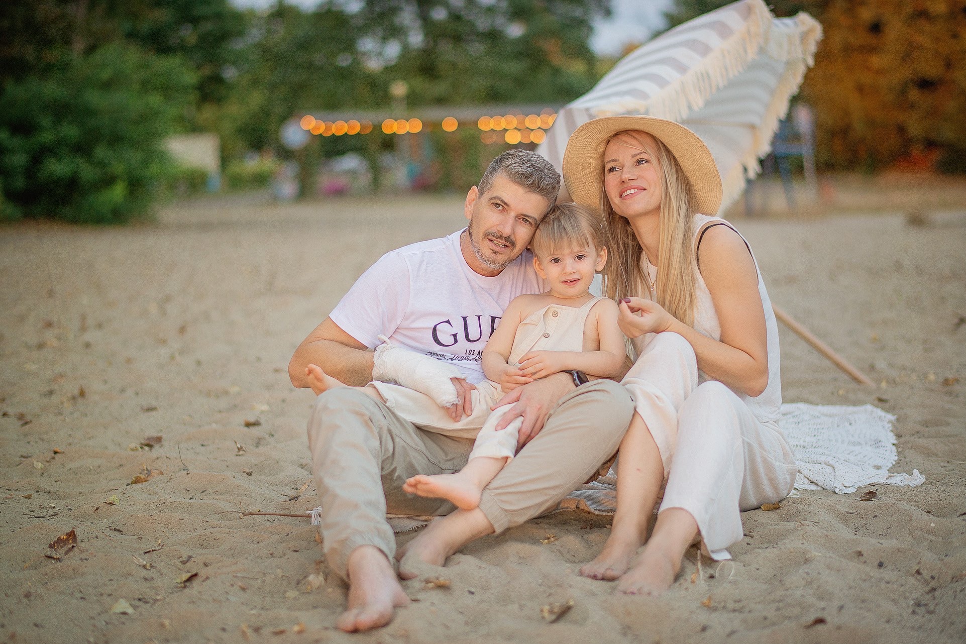 Rodzinne zdjęcie na plaży: rodzice z małym dzieckiem siedzą na piasku, w tle parasol i dekoracyjne lampki. Ojciec ma założony gips na ręce. Ubrani w jasne, letnie stroje.
