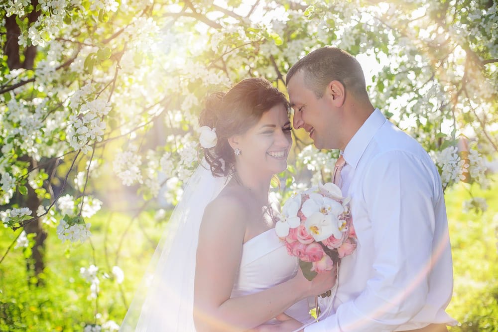 Uśmiechnięta para młoda, zbliżona twarzami, w otoczeniu kwitnących drzew w słoneczny dzień. Panna młoda trzyma bukiet różowych i białych kwiatów.