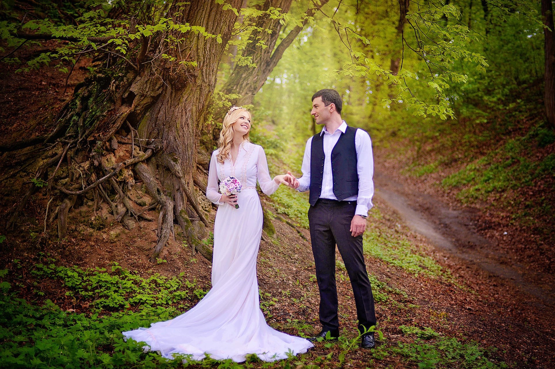 Para młoda trzymająca się za ręce w lesie, panna młoda w koronkowej sukni i z bukietem, pan młody w kamizelce, romantyczna sceneria z drzewami i zielenią.