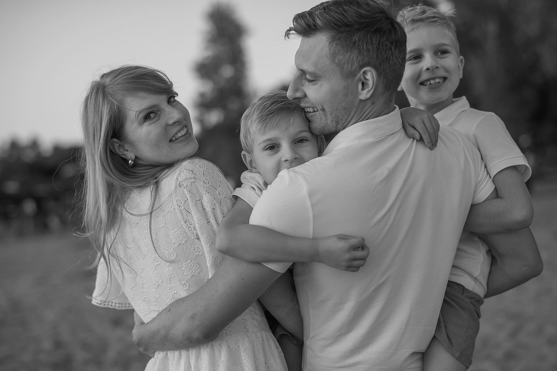 Czarno-białe, ciepłe ujęcie rodziny: mama w koronkowej sukience, tata i dwóch synów w objęciach, uśmiechnięci, na tle rozmytej zieleni. Naturalne światło.