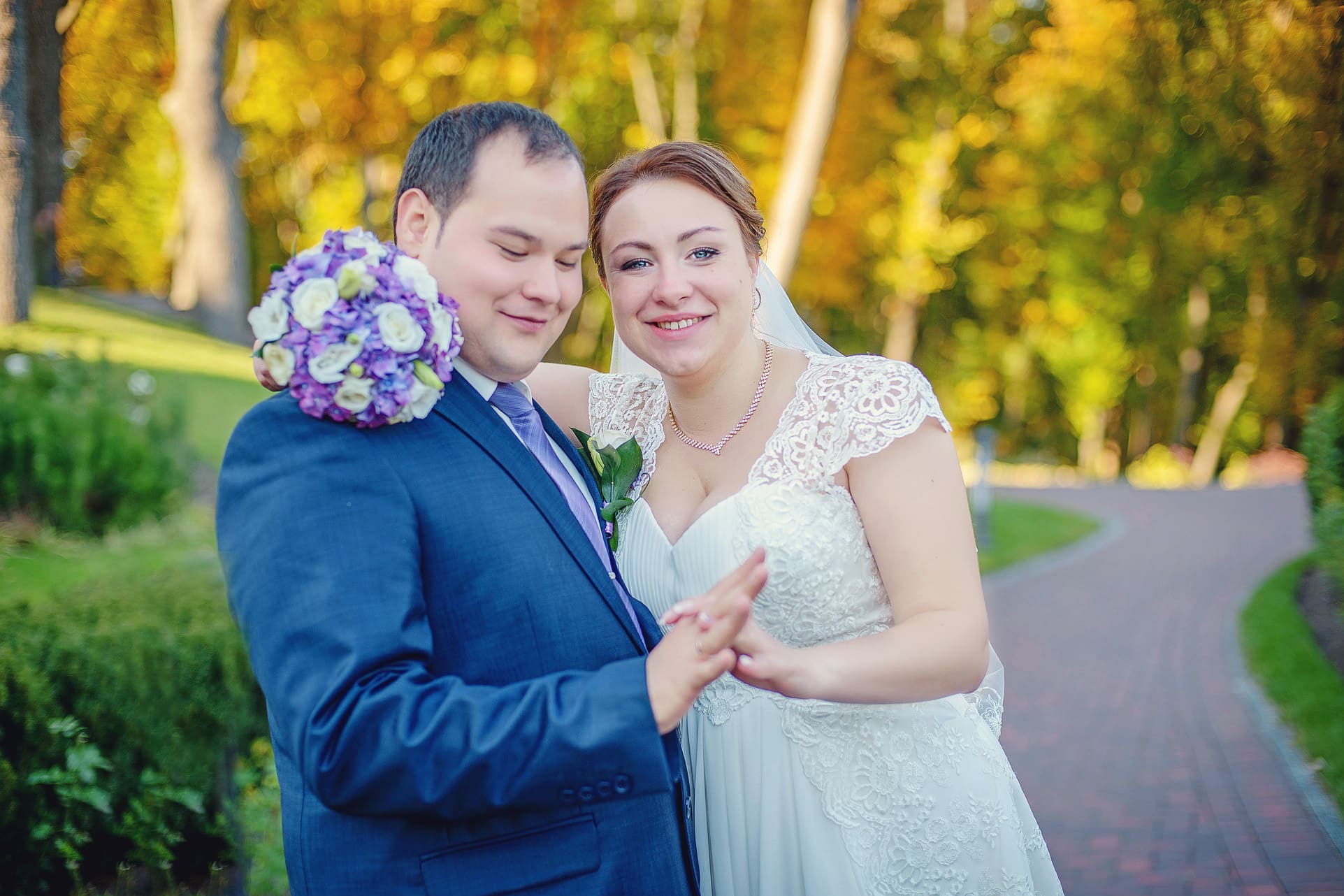Uśmiechnięta para młoda w objęciach na tle jesiennego parku. Panna młoda w koronkowej sukni, pan młody w granatowym garniturze z fioletowym krawatem.