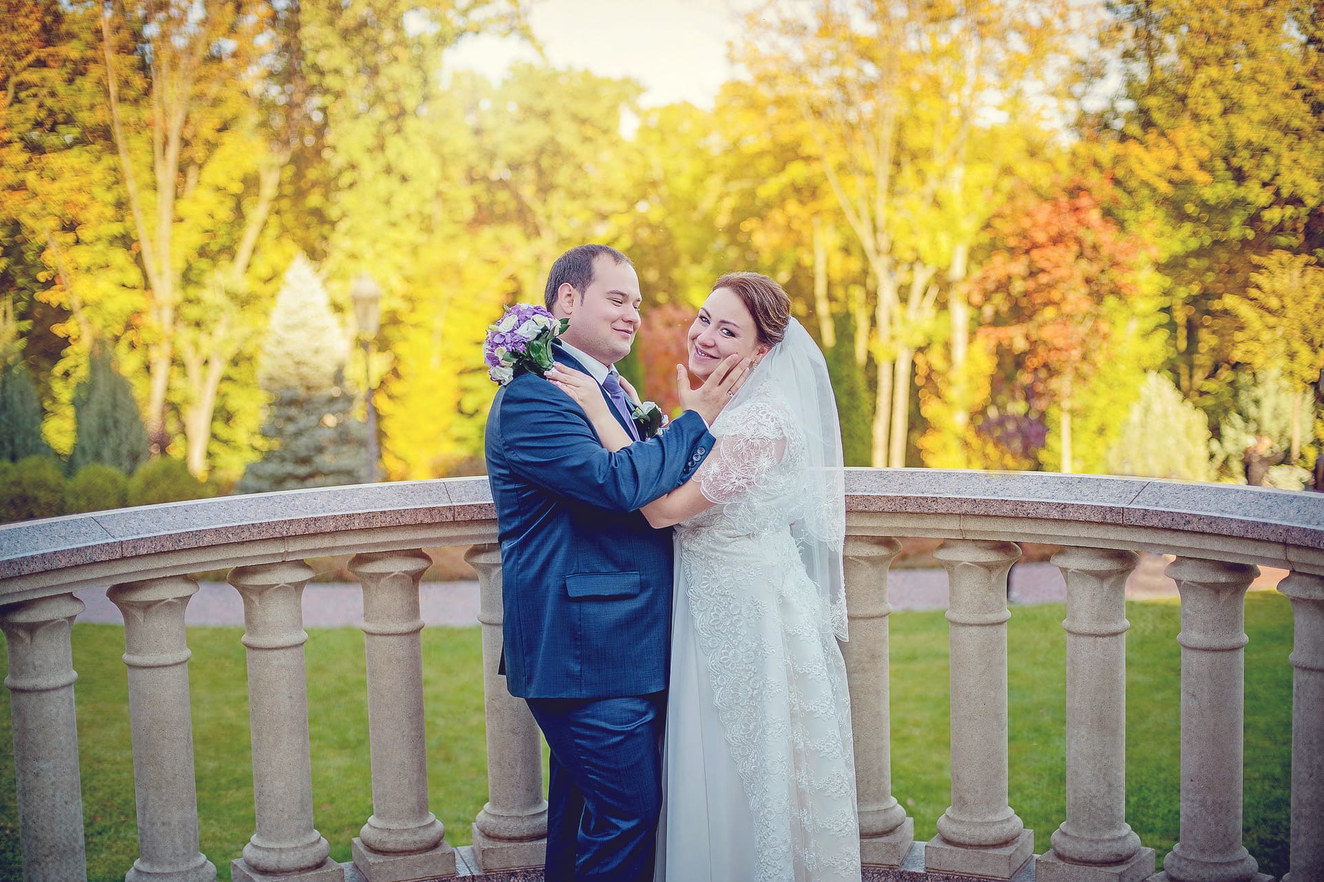 Uśmiechnięta para młoda w objęciach na tle jesiennego parku, on w granatowym garniturze, ona w koronkowej sukni ślubnej i welonie, trzyma bukiet.