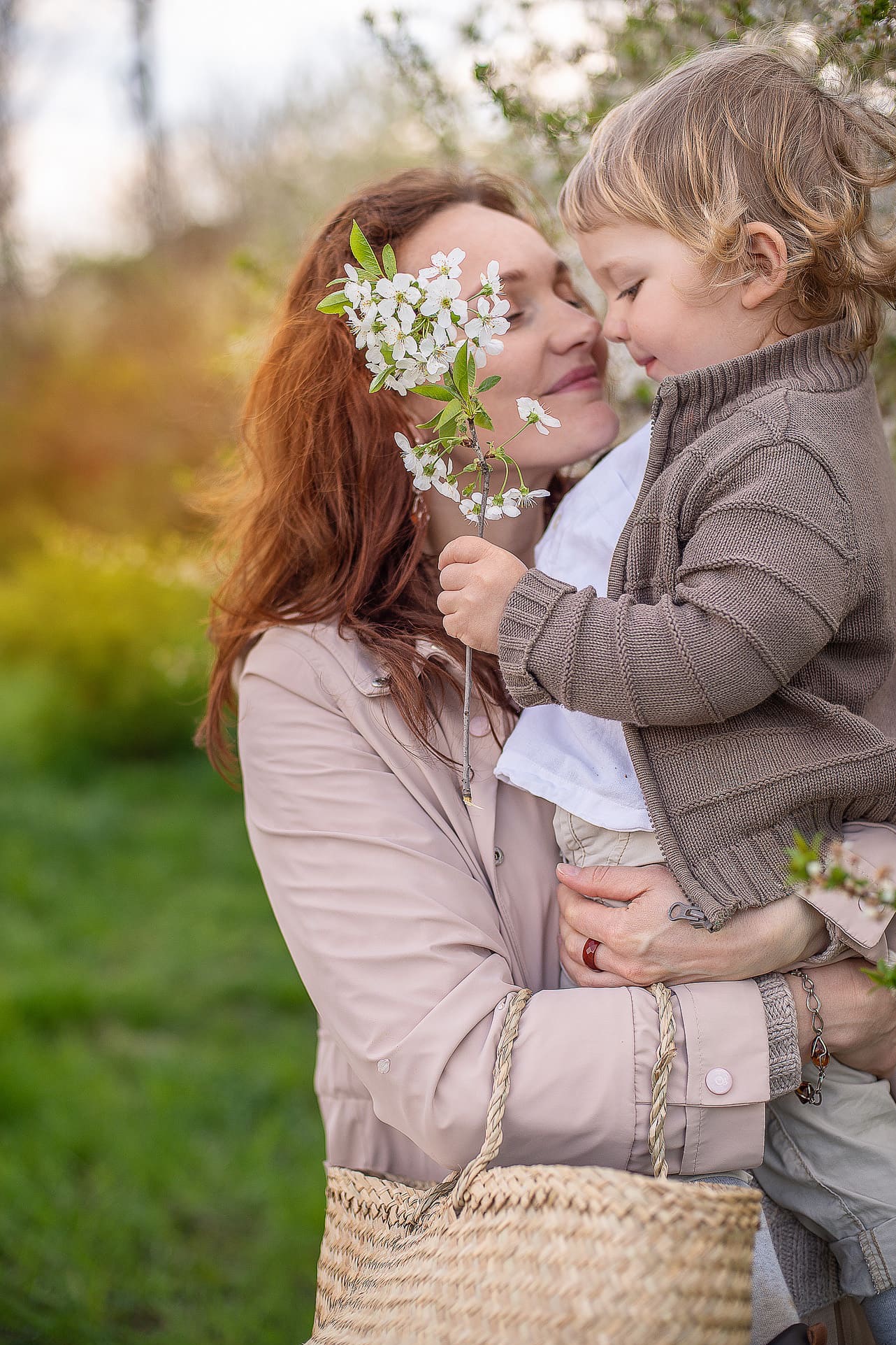 Uśmiechnięta matka z rudymi włosami trzyma synka z blond lokami, oboje dotykają się nosami, w tle kwitnące drzewa i koszyk.