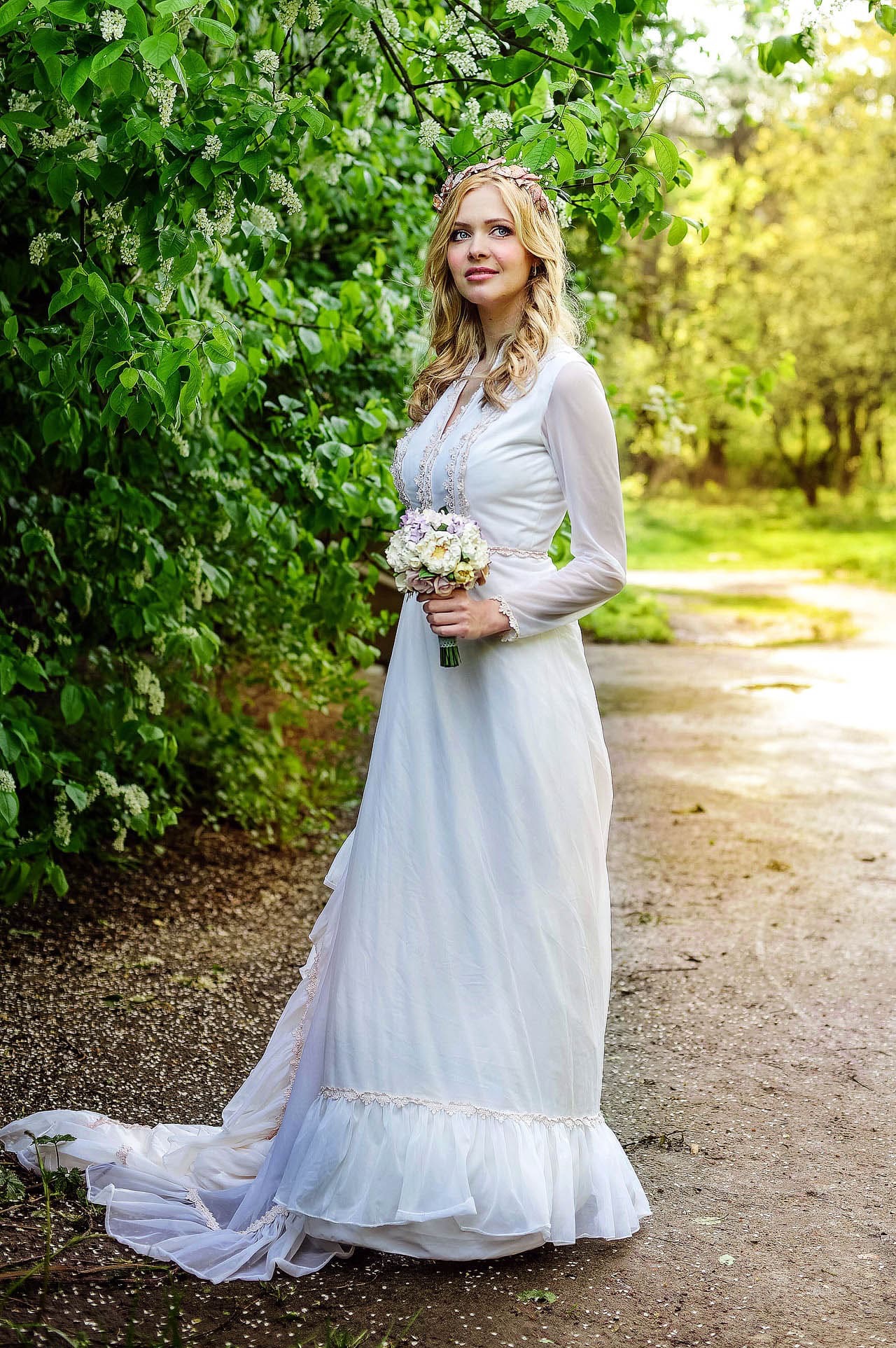 Panna młoda w białej sukni z bukietem, wianek na głowie, pozuje na tle kwitnących drzew w parku. Romantyczna sceneria, naturalne światło.