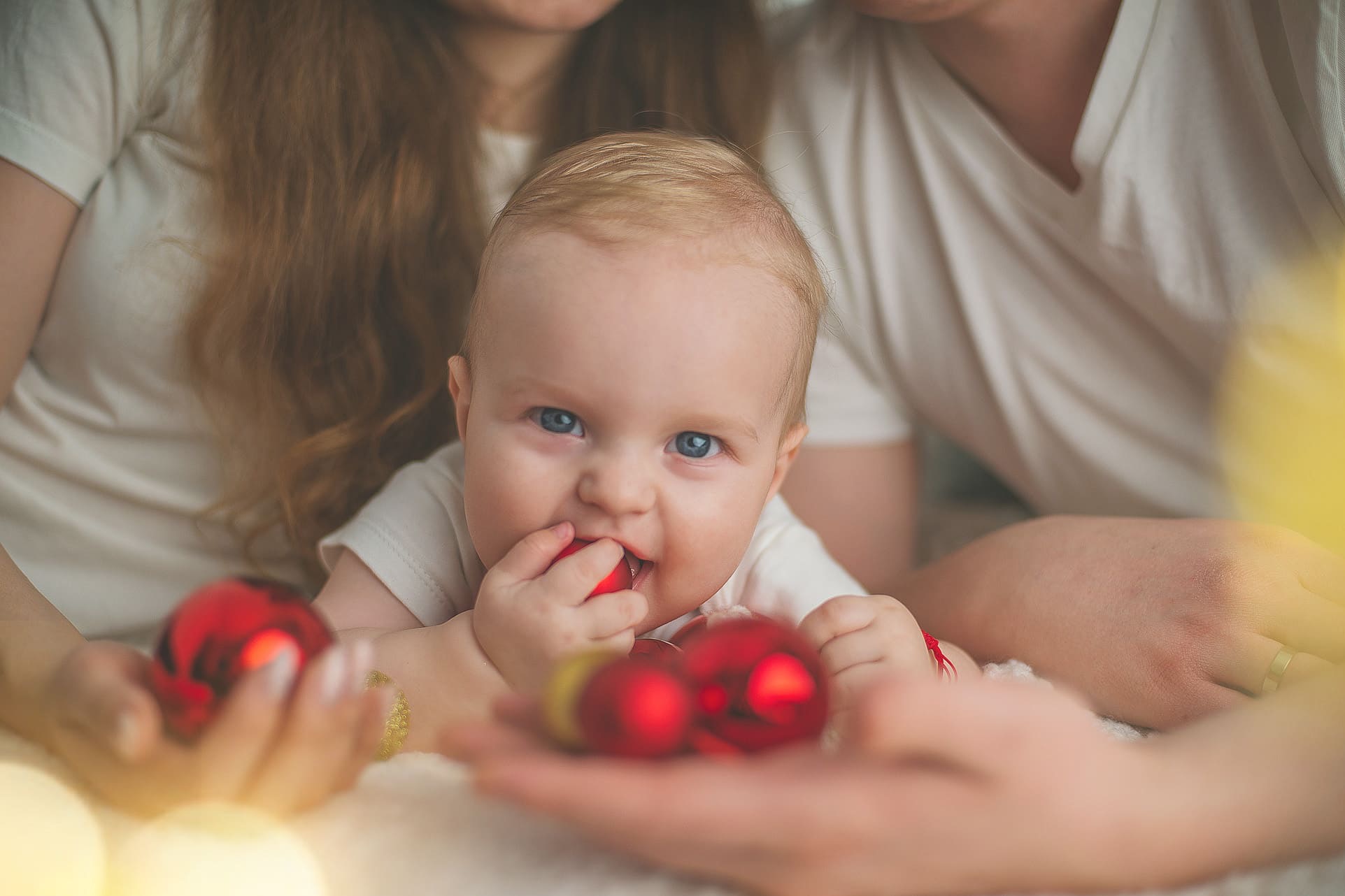 Uśmiechnięte niemowlę z niebieskimi oczami, trzymające czerwoną bombkę w ustach, w otoczeniu rodziców i świątecznych dekoracji. Ciepłe, rozproszone światło.