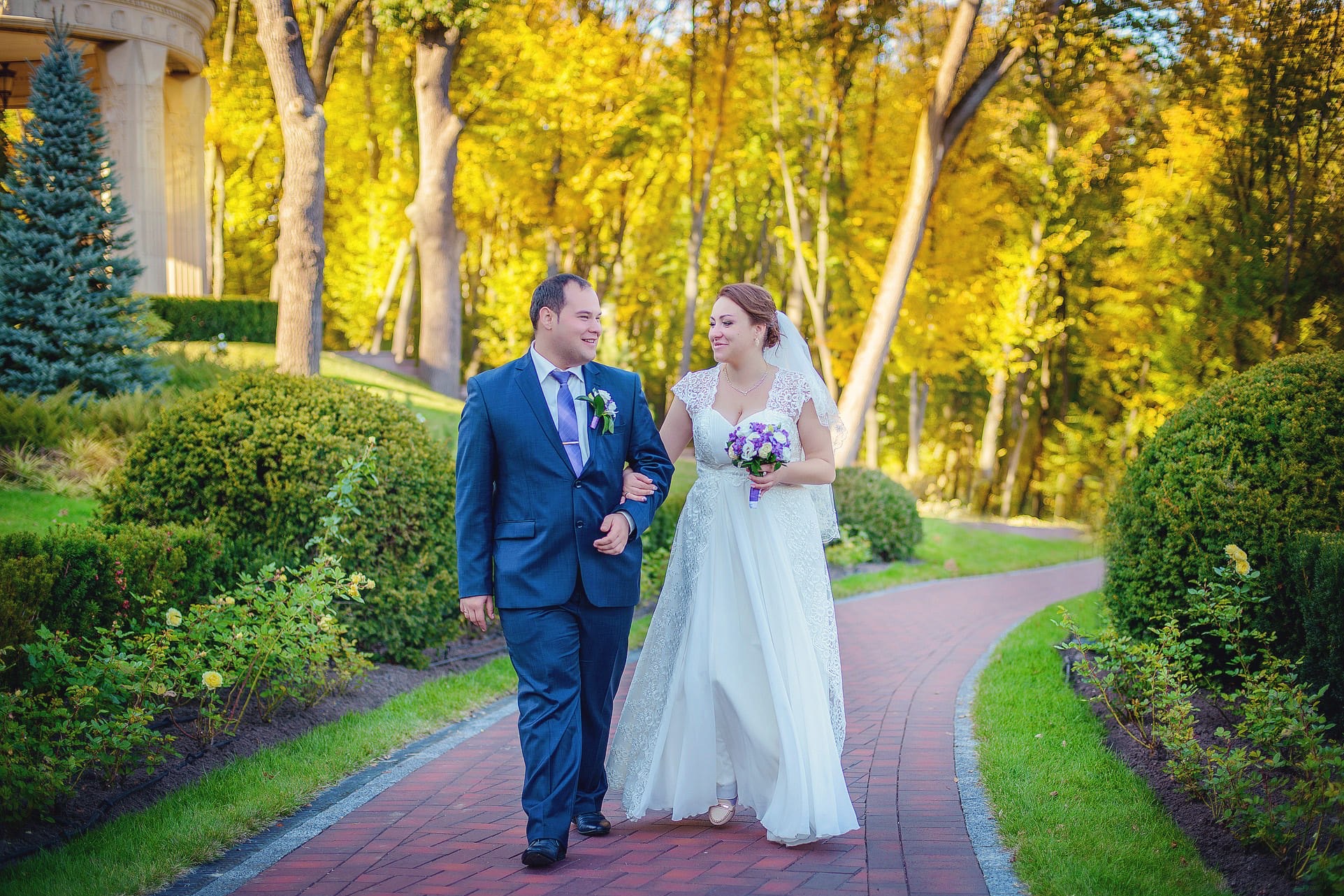 Para młoda, on w granatowym garniturze z fioletowym krawatem, ona w białej sukni ślubnej z bukietem, spacerują po czerwonej ścieżce w parku w słoneczny dzień.