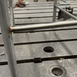 Detal spawanej konstrukcji metalowej z rur na stole warsztatowym; widoczne spawy, otwory w blacie i rękawice robocze w tle. Surowy, industrialny charakter.