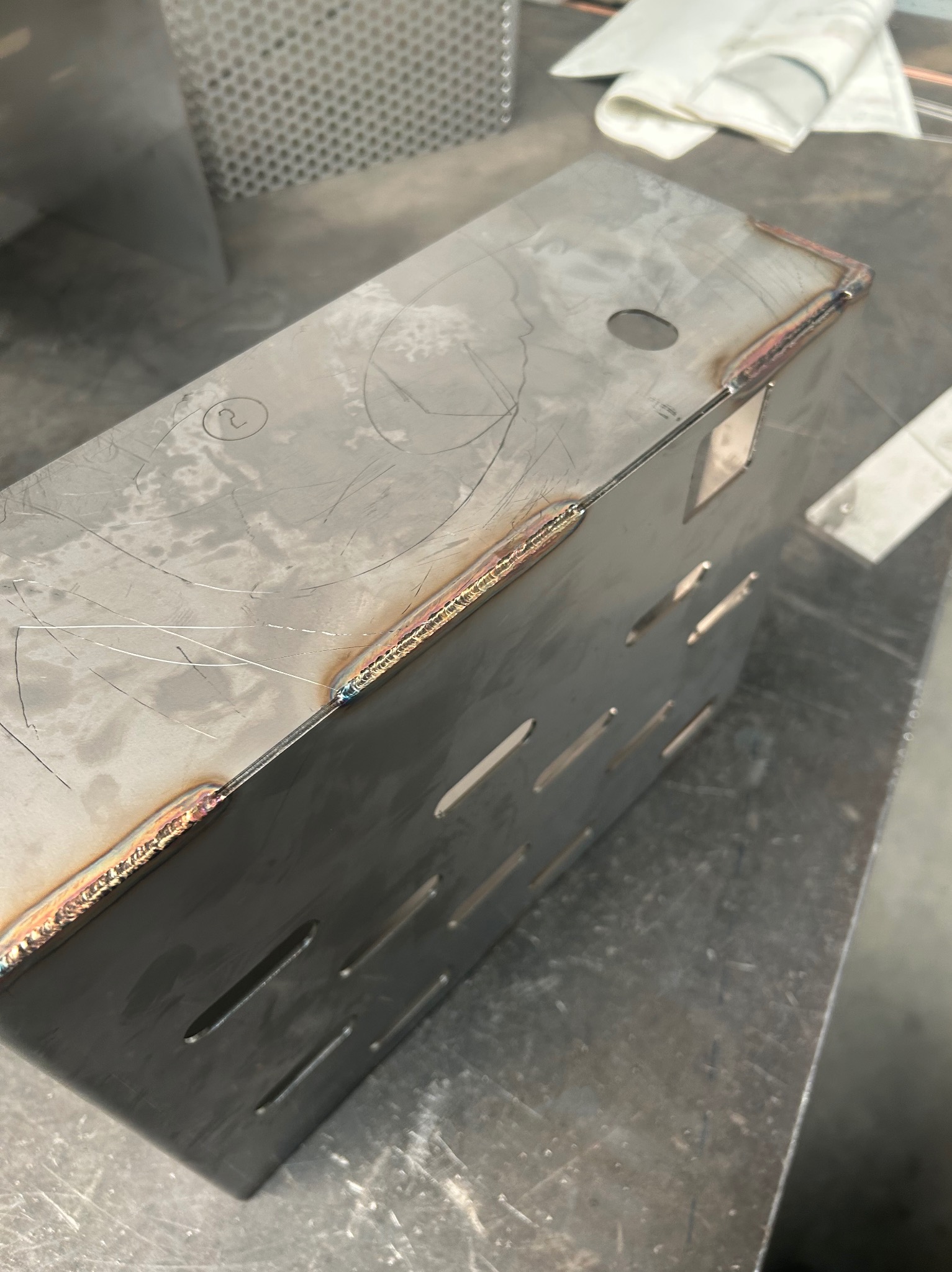 Spawana, metalowa obudowa z otworami wentylacyjnymi, widoczne spawy i ślady obróbki. Obudowa stoi na metalowym stole warsztatowym.