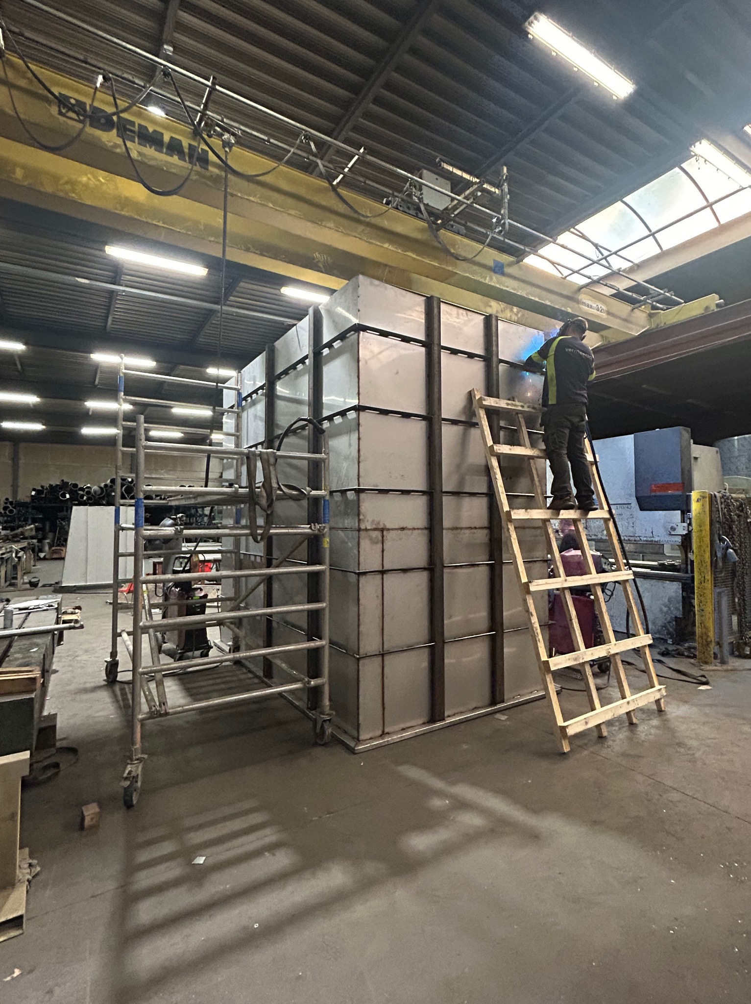 Spawacz na drabinie łączy metalowe elementy konstrukcji w hali. Widoczne rusztowanie i suwnica. Iskry podczas spawania.