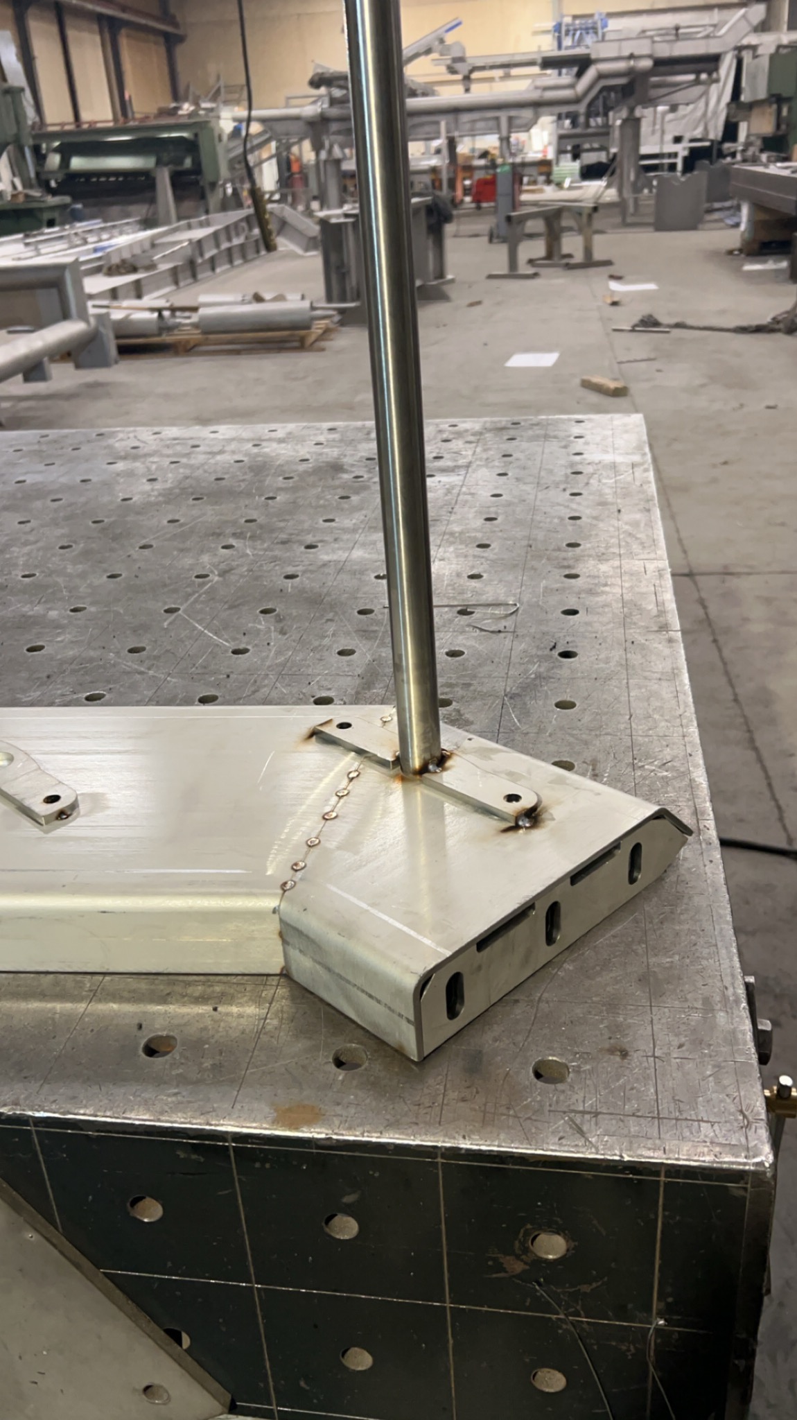 Szlifowany element metalowy z przymocowanym słupkiem, spawany na perforowanym stole warsztatowym w hali produkcyjnej. Widoczne ślady spawania i obróbki.