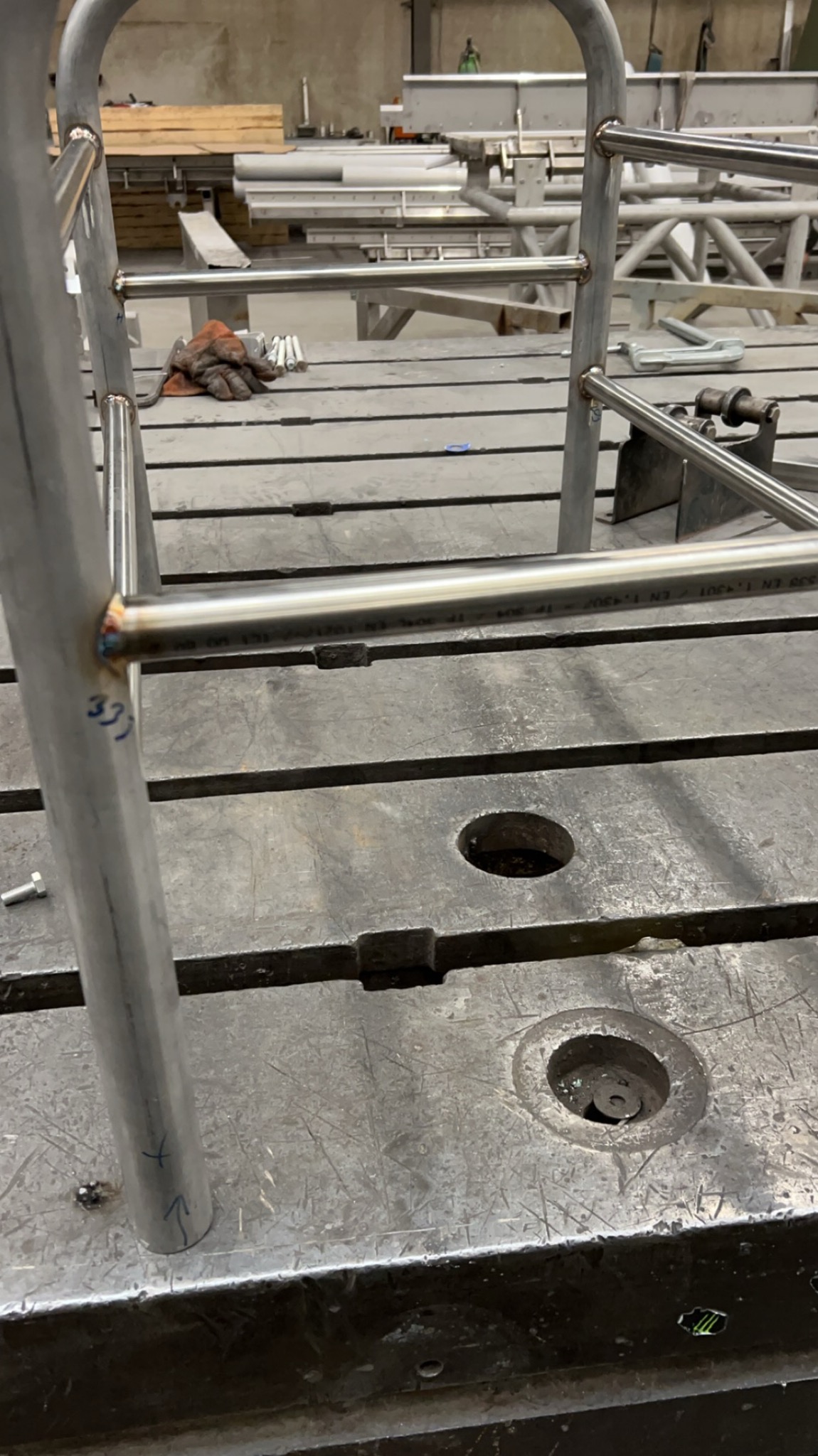 Detal spawanej konstrukcji metalowej z rur na stole warsztatowym; widoczne spawy, otwory w blacie i rękawice robocze w tle. Surowy, industrialny charakter.