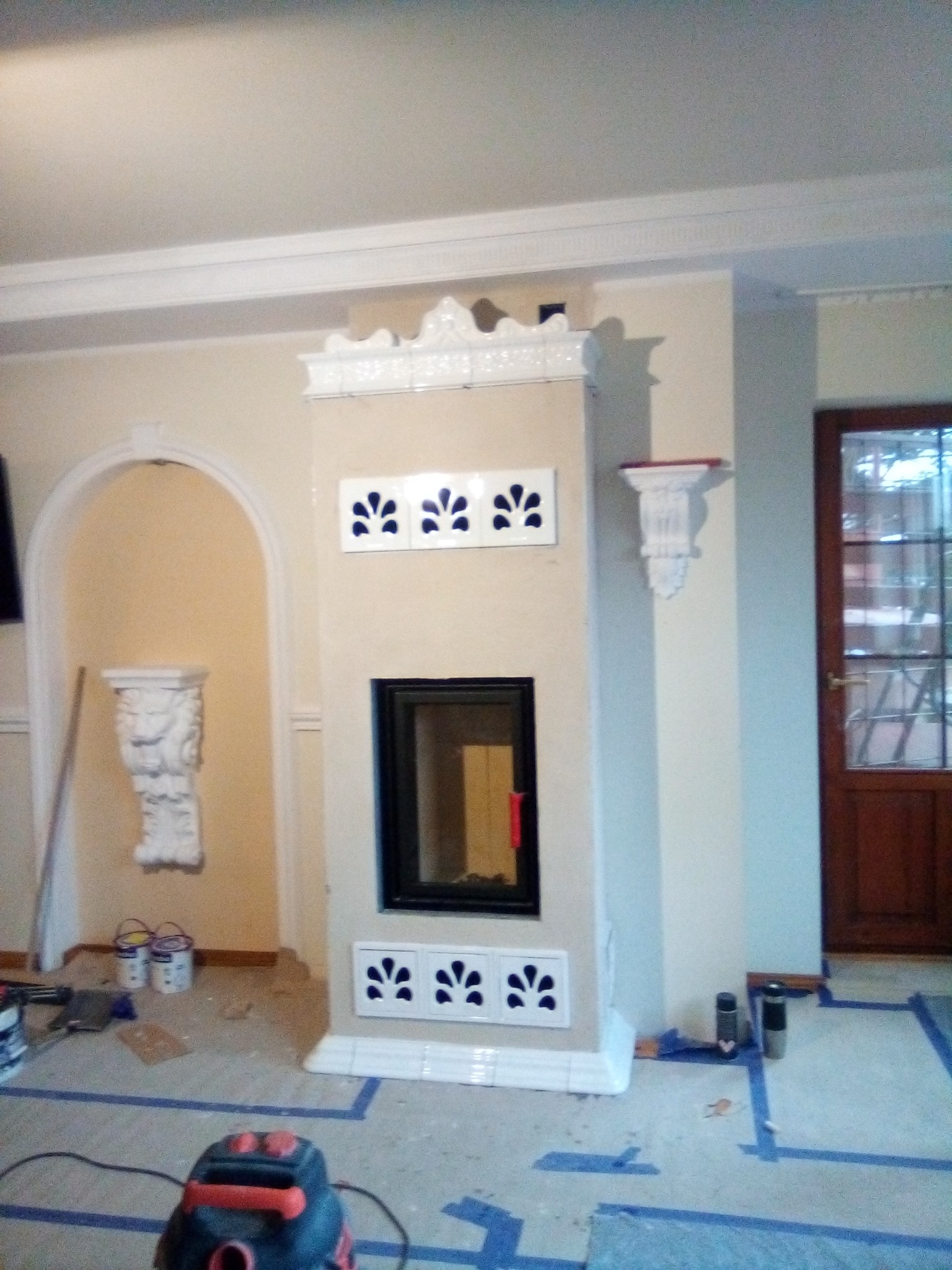 Wnętrze w trakcie prac wykończeniowych z widocznym kominkiem obłożonym kremowymi płytkami z dekoracyjnymi, niebieskimi wstawkami, zabezpieczona podłoga taśmą malarską, oraz odkurzacz budowlany.