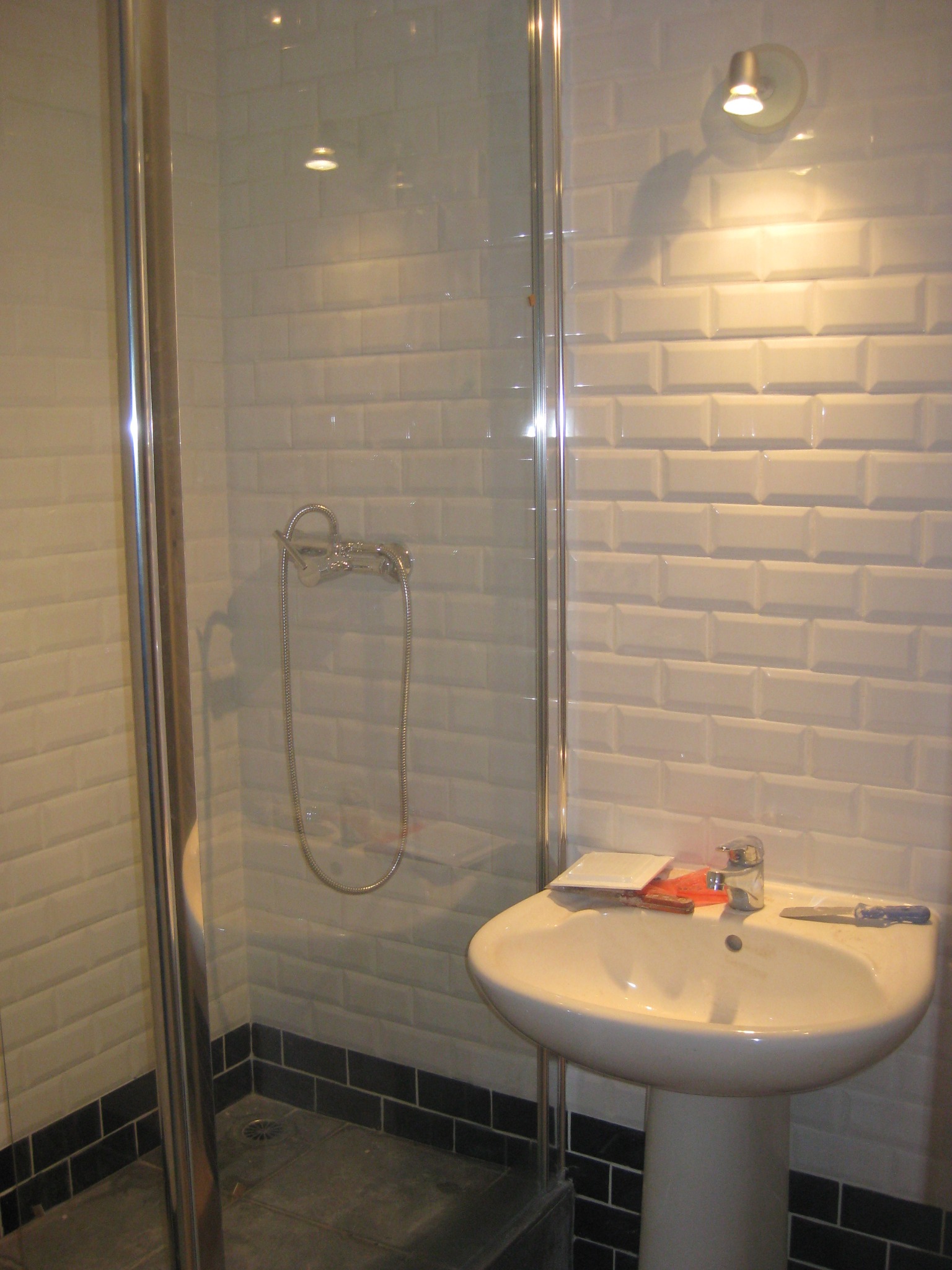 Umywalka na postumencie z baterią i akcesoriami glazurniczymi obok kabiny prysznicowej wyłożonej białymi, cegiełkowymi płytkami, dolny pas wykończony czarną glazurą.