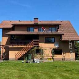 SZERSZEŃ Dawid Leśniowski - Pracownik w czerwonej koszulce na dachu domu z brązową dachówką, prawdopodobnie podczas montażu okna dachowego, z widocznymi narzędziami i wiadrami z farbą na trawniku poniżej.