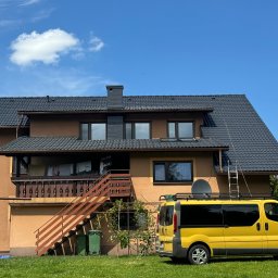 SZERSZEŃ Dawid Leśniowski - Dwupiętrowy dom z ciemnym dachem, balkonem i drewnianymi schodami zewnętrznymi, z żółtym busem zaparkowanym na trawniku przed domem. Na dachu widoczna antena satelitarna i drabina oparta o rynnę.