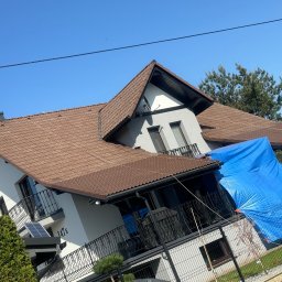 SZERSZEŃ Dawid Leśniowski - Dom jednorodzinny z brązowym dachem krytym dachówką, zabezpieczony niebieską plandeką na części balkonu, widok w słoneczny dzień.