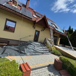 SZERSZEŃ Dawid Leśniowski - Montaż paneli fotowoltaicznych na dachu domu z dachówką ceramiczną, zabezpieczonym folią, widoczne drabiny i pracownik w czerwonej koszulce.