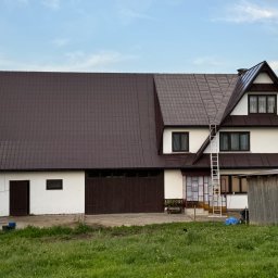 SZERSZEŃ Dawid Leśniowski - Dwukondygnacyjny dom z brązowym dachem z blachy trapezowej, częściowo odnowionym, z drabiną opartą o ścianę, widok z poziomu trawnika.