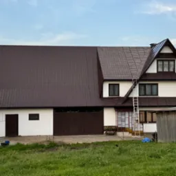 Dwukondygnacyjny dom z brązowym dachem z blachy trapezowej, częściowo odnowionym, z drabiną opartą o ścianę, widok z poziomu trawnika.
