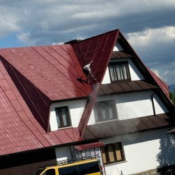SZERSZEŃ Dawid Leśniowski - Pracownik zabezpieczony linami, czyści dach domu z czerwonej dachówki za pomocą myjki ciśnieniowej, widoczny żółty bus firmy na tle białej elewacji.