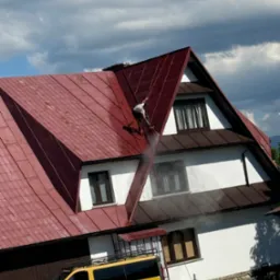 Pracownik zabezpieczony linami, czyści dach domu z czerwonej dachówki za pomocą myjki ciśnieniowej, widoczny żółty bus firmy na tle białej elewacji.