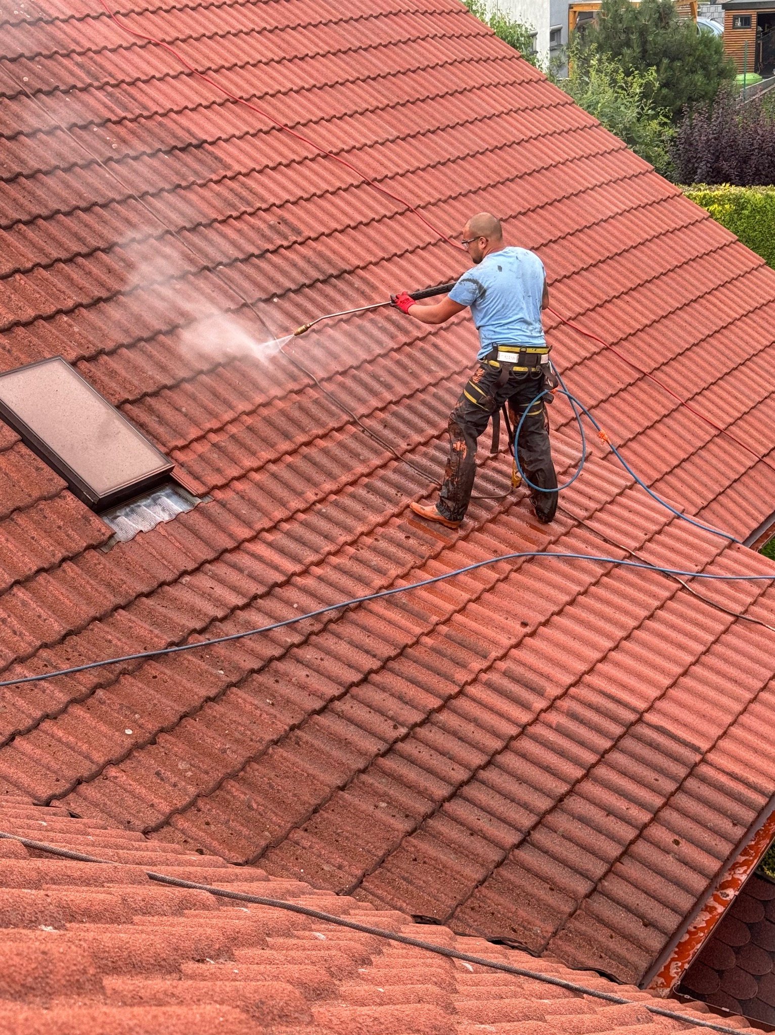 Pracownik w czerwonych rękawicach czyści czerwoną dachówkę za pomocą myjki ciśnieniowej, widoczna para wodna i częściowo oczyszczona powierzchnia dachu z oknem dachowym.