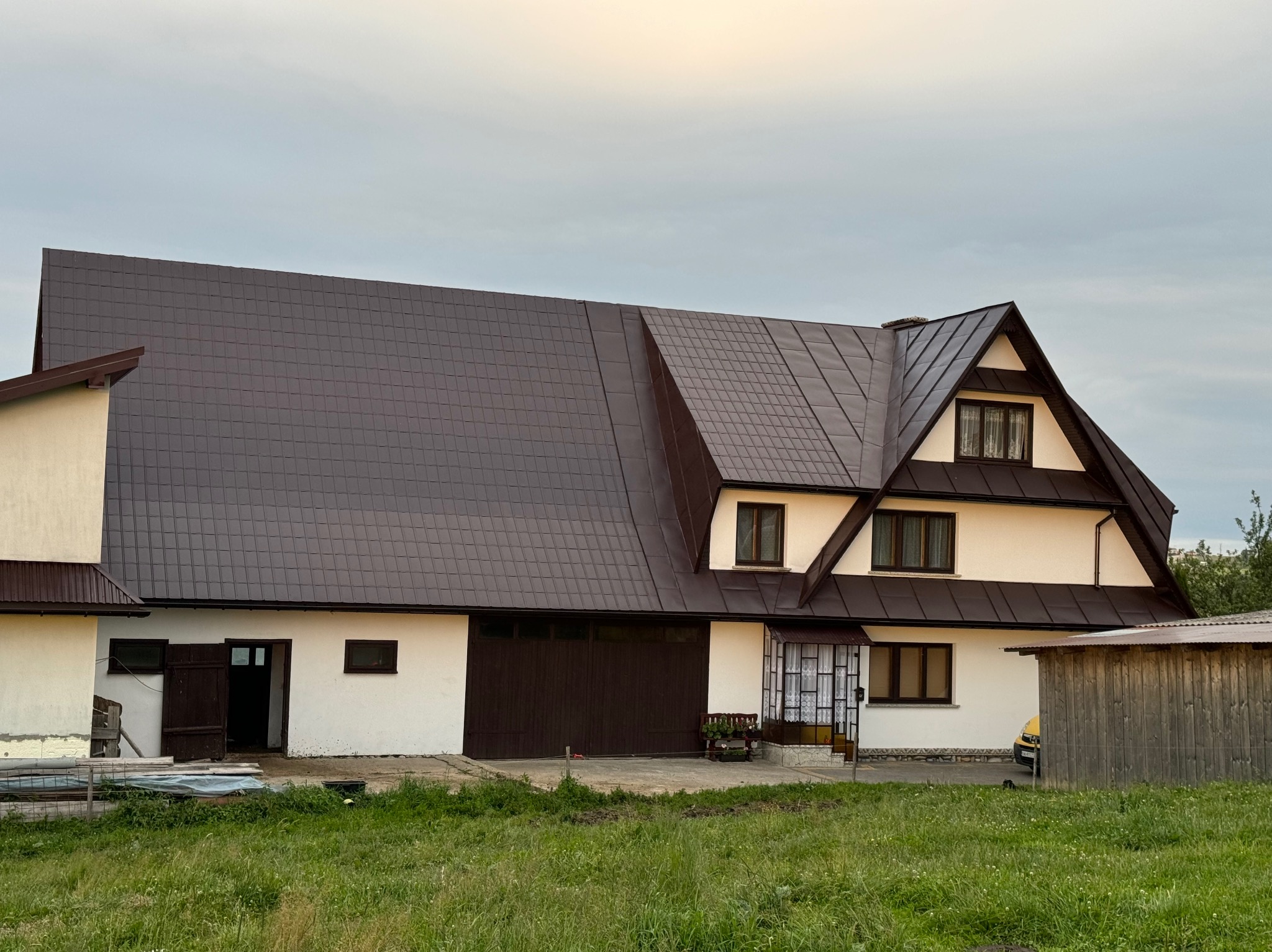 Dwukondygnacyjny budynek z nowym, brązowym dachem z blachodachówki, białą elewacją i brązowymi elementami wykończeniowymi, widoczny z poziomu trawnika.