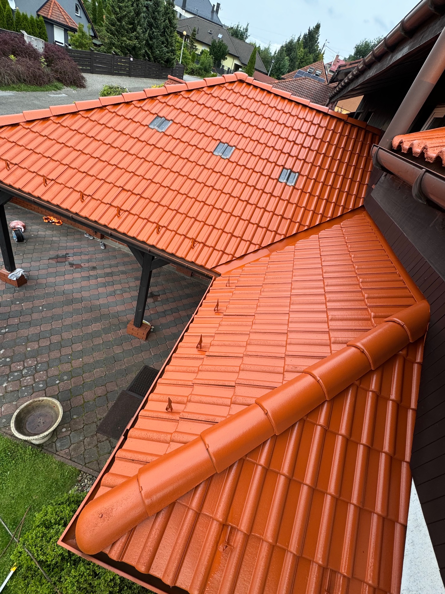 Świeżo pomalowany dach z dachówki ceramicznej w odcieniach terakoty, widok z góry, z widocznymi elementami rynny i fragmentem podwórza z kostki brukowej.