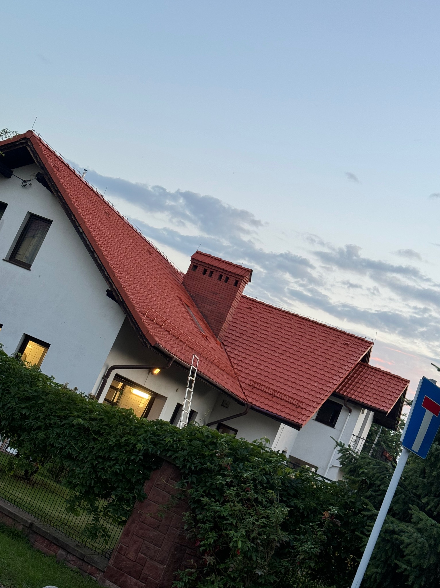 Dom jednorodzinny z czerwonym dachem z dachówki ceramicznej, widoczna drabina oparta o rynnę, w tle błękitne niebo z chmurami.