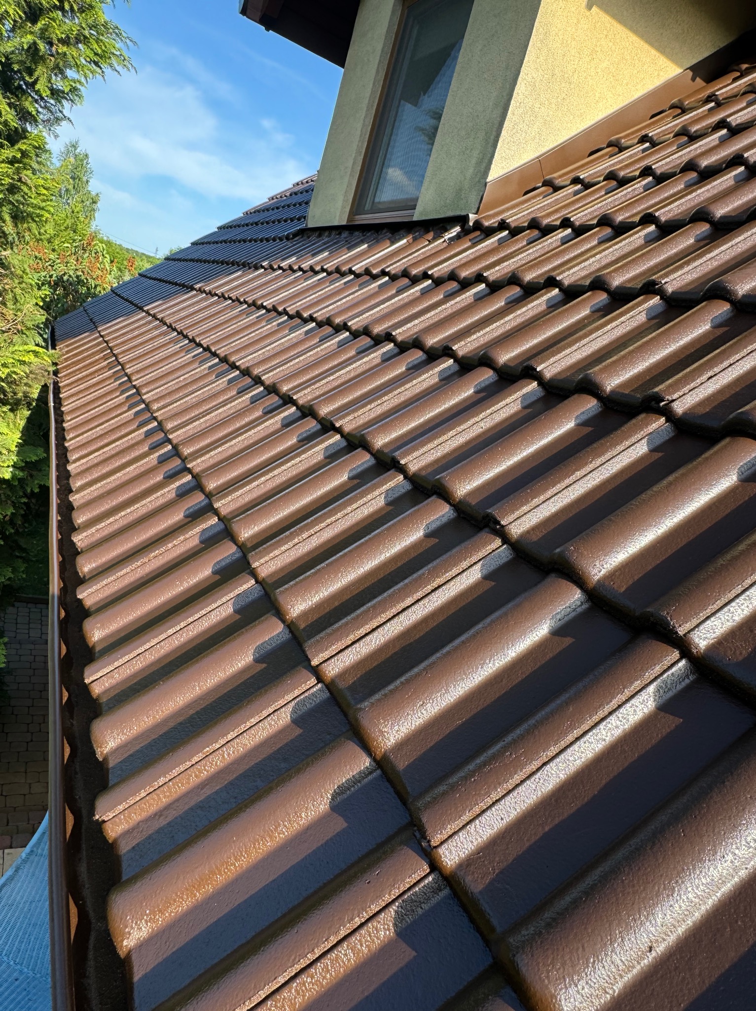 Błyszczący, brązowy dach z dachówki ceramicznej, świeżo po renowacji, widok z góry w słoneczny dzień, z fragmentem okna dachowego i zieleni w tle.