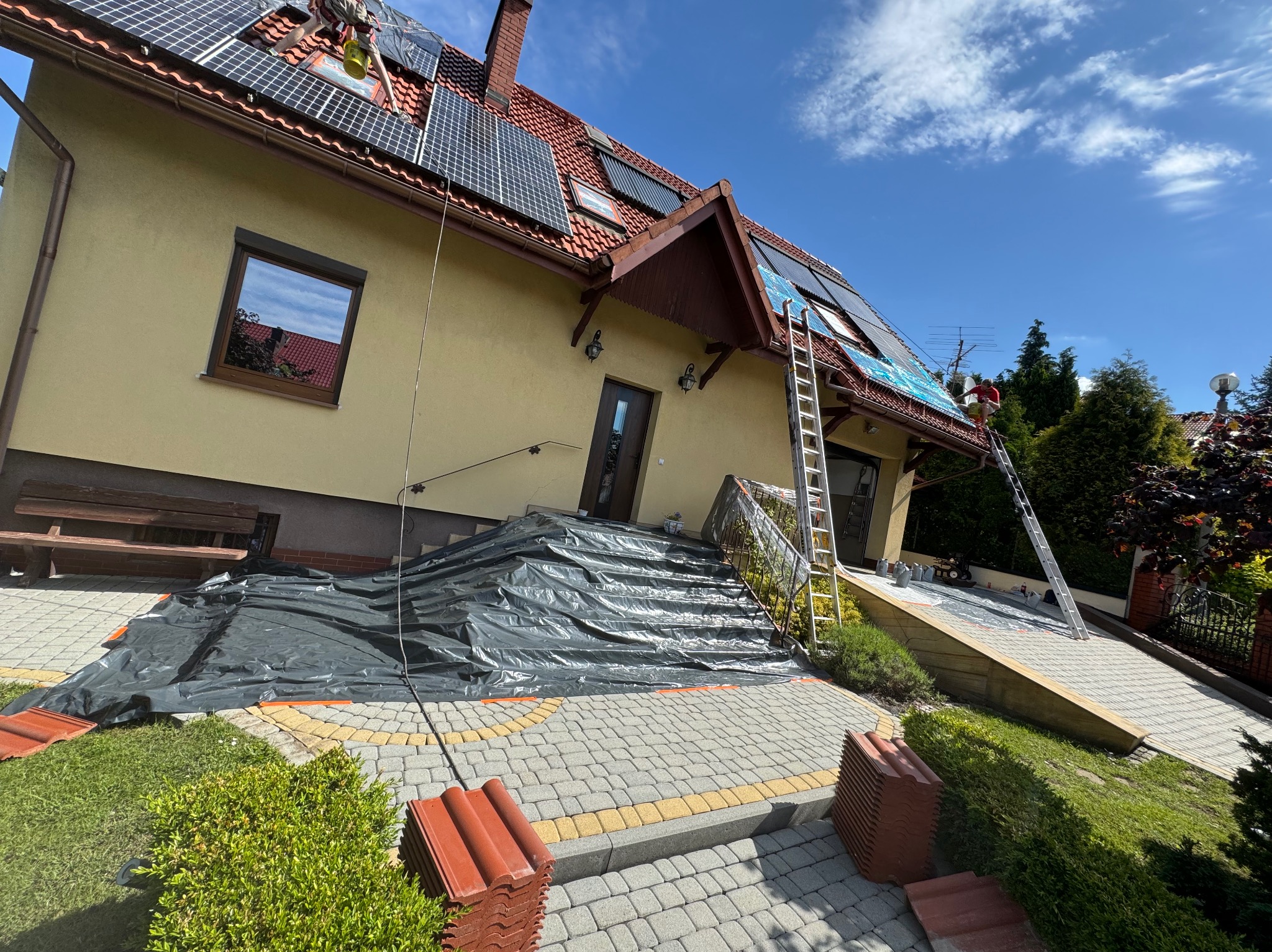Montaż paneli fotowoltaicznych na dachu domu z dachówką ceramiczną, zabezpieczonym folią, widoczne drabiny i pracownik w czerwonej koszulce.