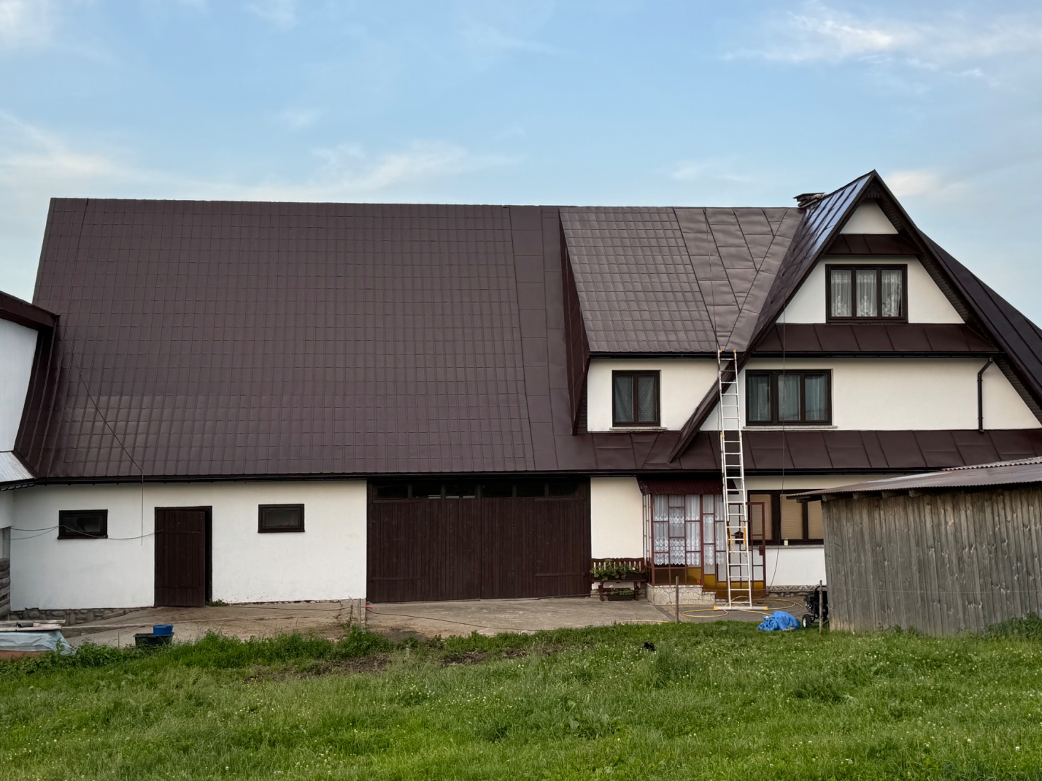 Dwukondygnacyjny dom z brązowym dachem z blachy trapezowej, częściowo odnowionym, z drabiną opartą o ścianę, widok z poziomu trawnika.