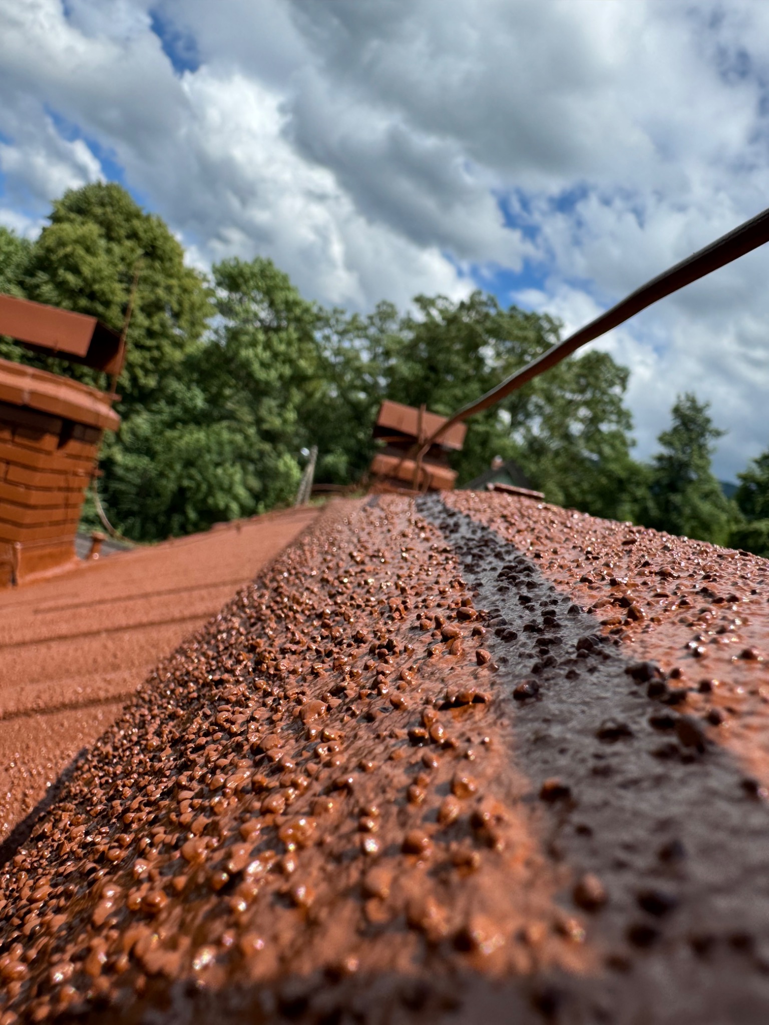 Pomarańczowo-brązowy dach pokryty drobnym granulatem, widok z bliska na strukturę powierzchni, w tle zieleń drzew i błękitne niebo z chmurami.