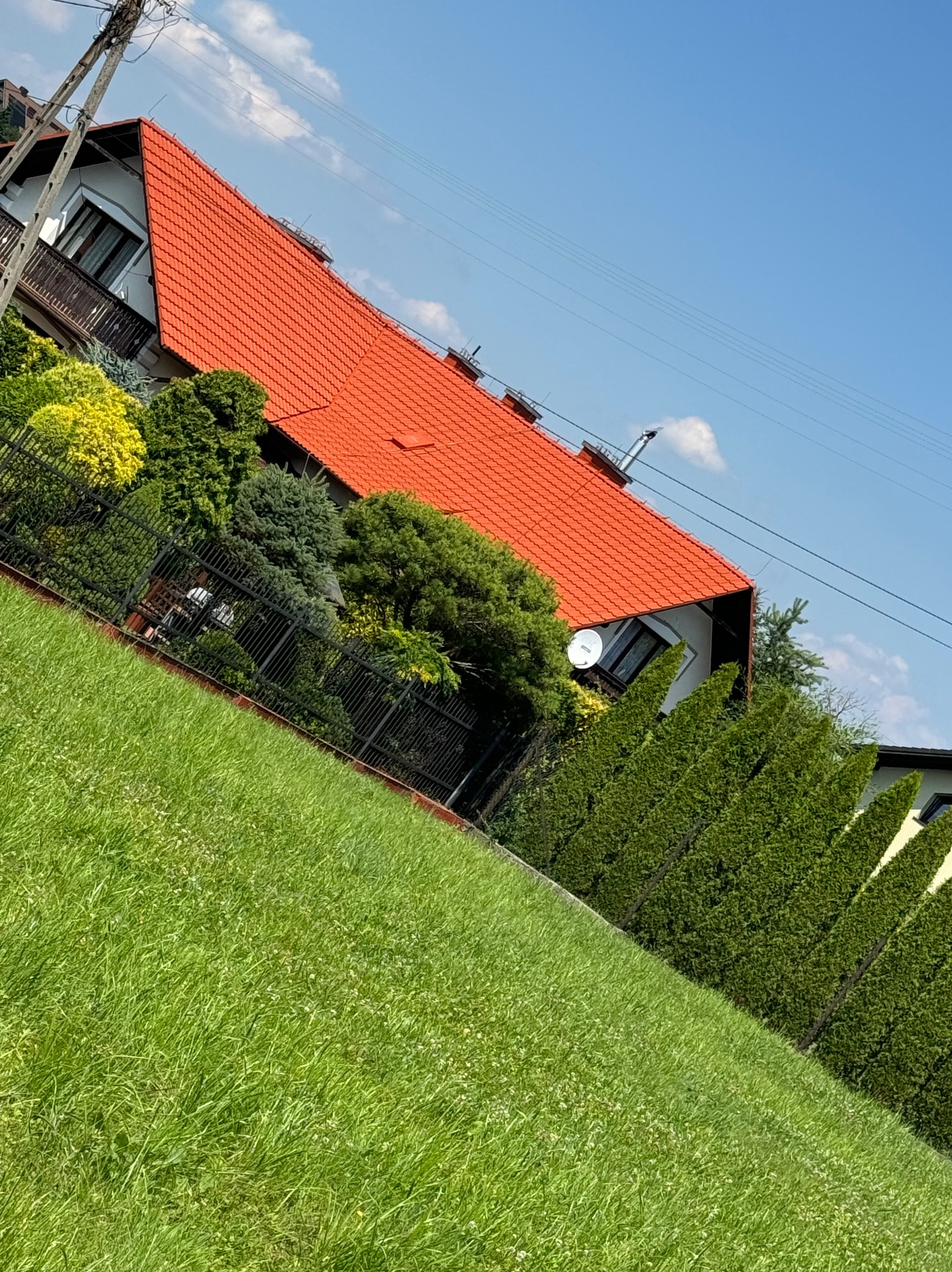 Dom jednorodzinny z intensywnie pomarańczowym dachem z dachówki ceramicznej, widoczny z poziomu trawnika, z anteną satelitarną na ścianie i kominem, na tle błękitnego nieba.