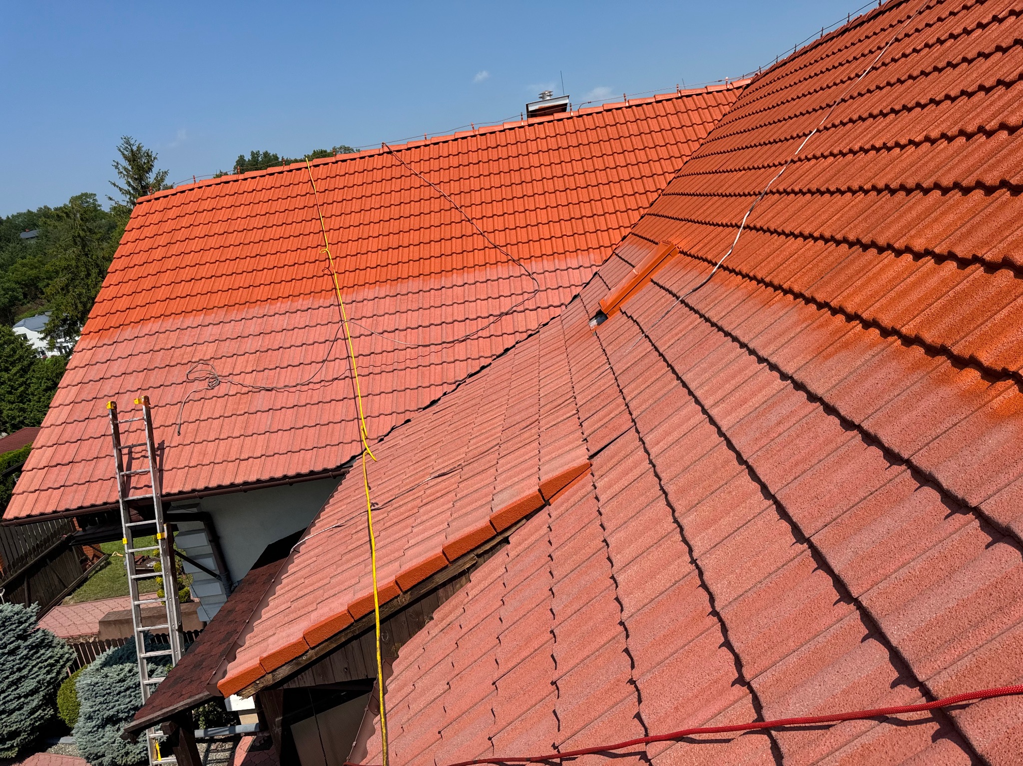 Częściowo pomalowany, czerwony dach z dachówki ceramicznej, widoczna drabina oparta o budynek i żółty kabel na dachu, błękitne niebo w tle.