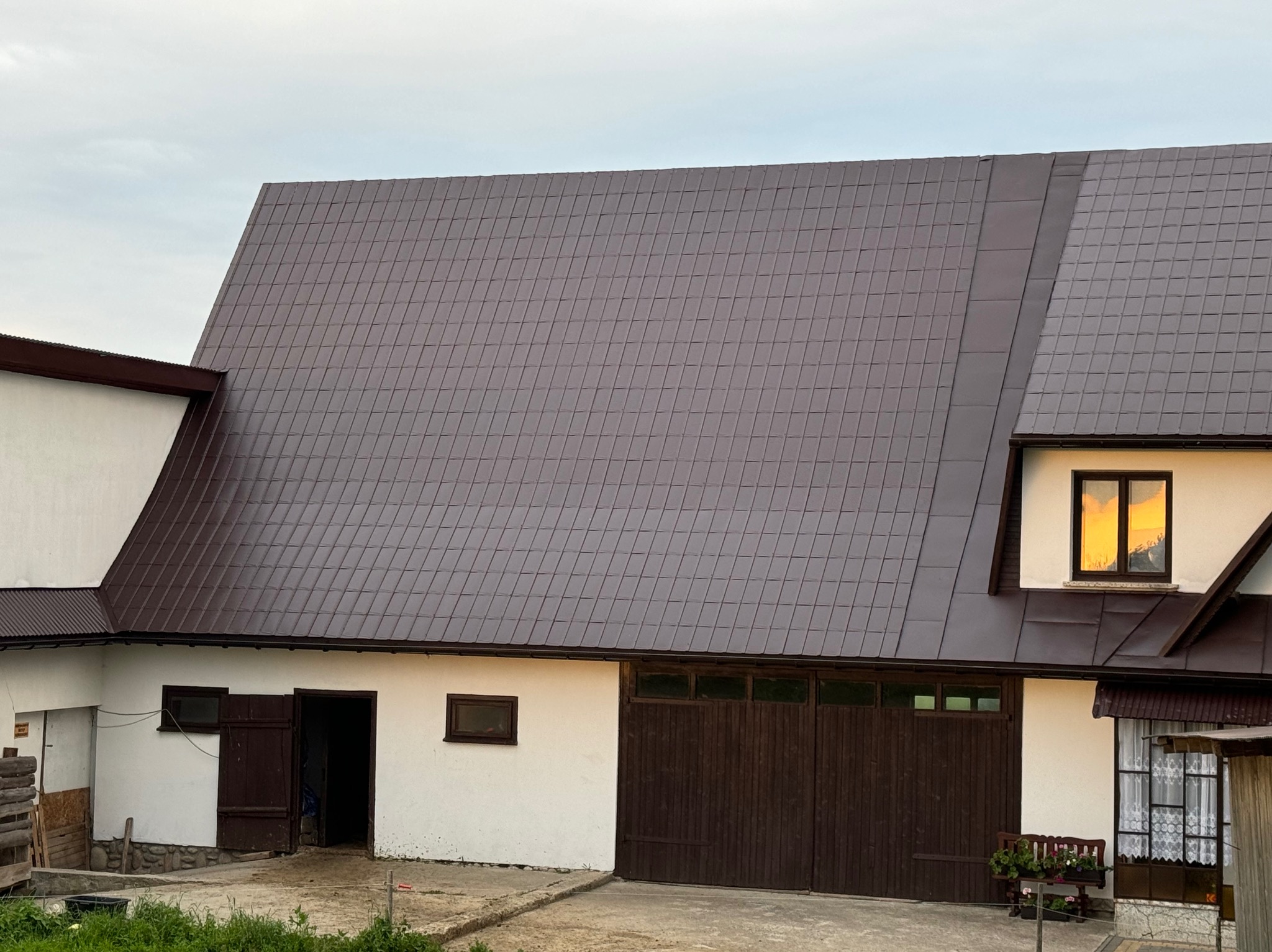 Brązowy dach z blachy trapezowej na budynku gospodarczym z białymi ścianami i drewnianymi wrotami garażowymi.